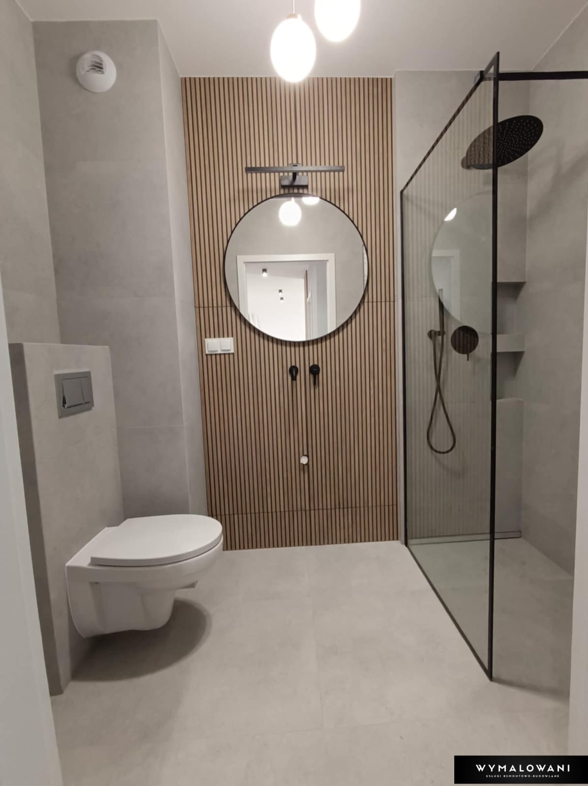 Nowoczesna łazienka z kabiną prysznicową typu walk-in, okrągłym lustrem z podświetleniem i drewnianą ścianą z pionowych paneli, wisząca toaleta, szara płytka podłogowa i ścienna.