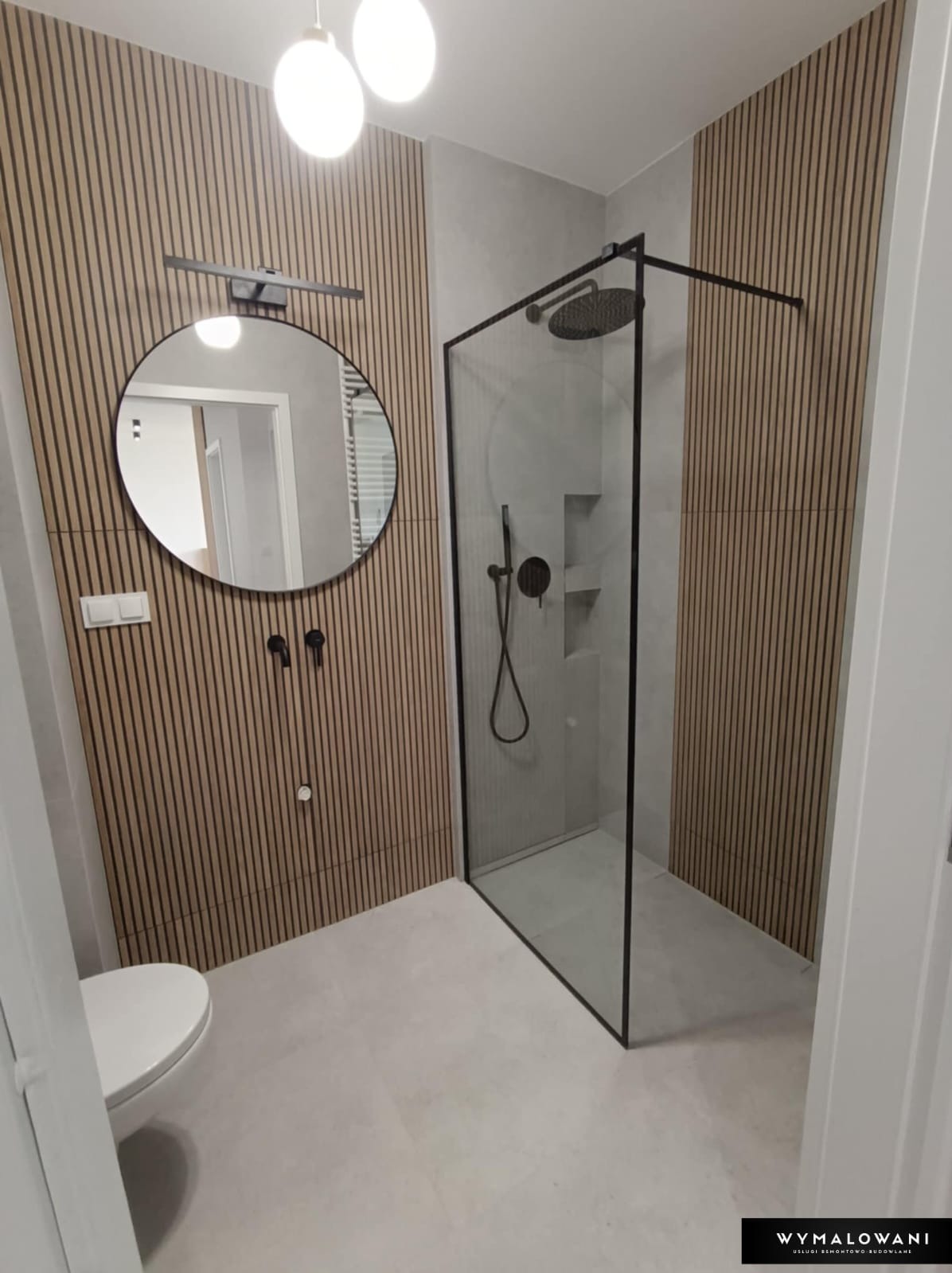 Nowoczesna łazienka po remoncie z kabiną prysznicową typu walk-in z czarnymi profilami, drewnianymi lamelami na ścianie, okrągłym lustrem z podświetleniem i minimalistyczną armaturą w kolorze czarnym.