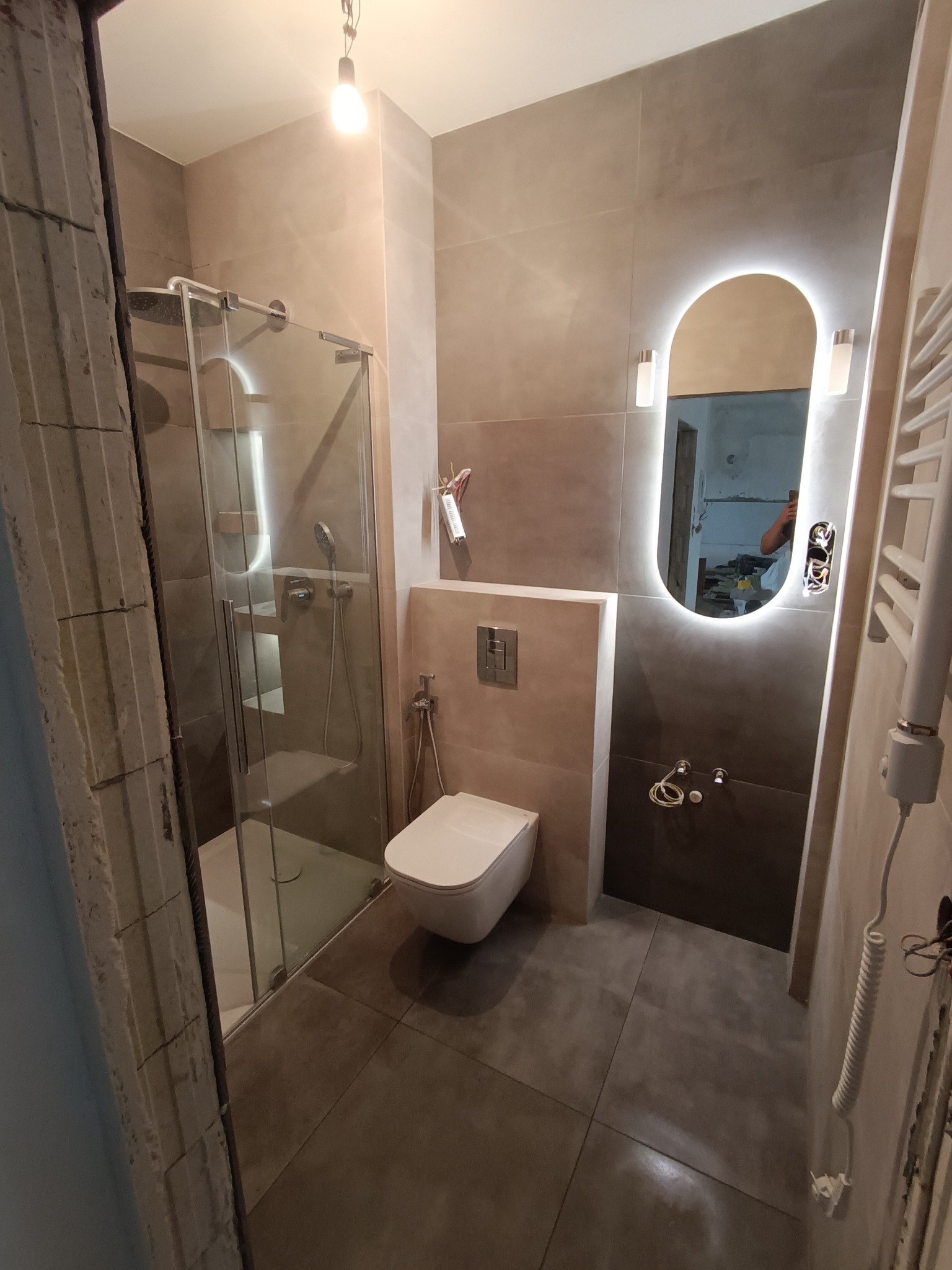 Nowoczesna łazienka w odcieniach szarości, z kabiną prysznicową, podświetlanym lustrem i wiszącą toaletą, widok na niedokończone obramowanie drzwi.