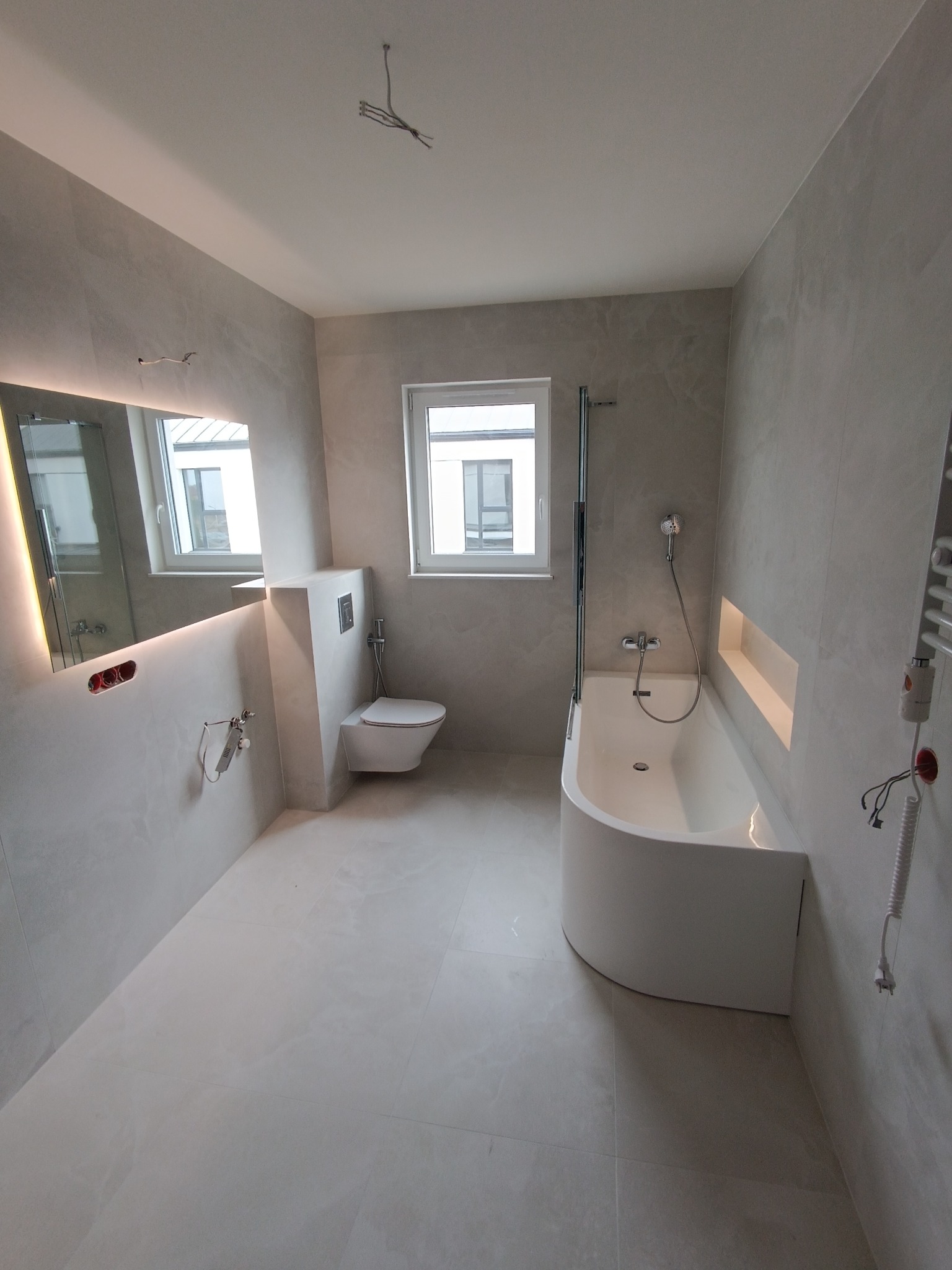 Jasna łazienka z wanną, podwieszaną toaletą i podświetlanym lustrem, widoczne niedokończone podłączenia elektryczne.