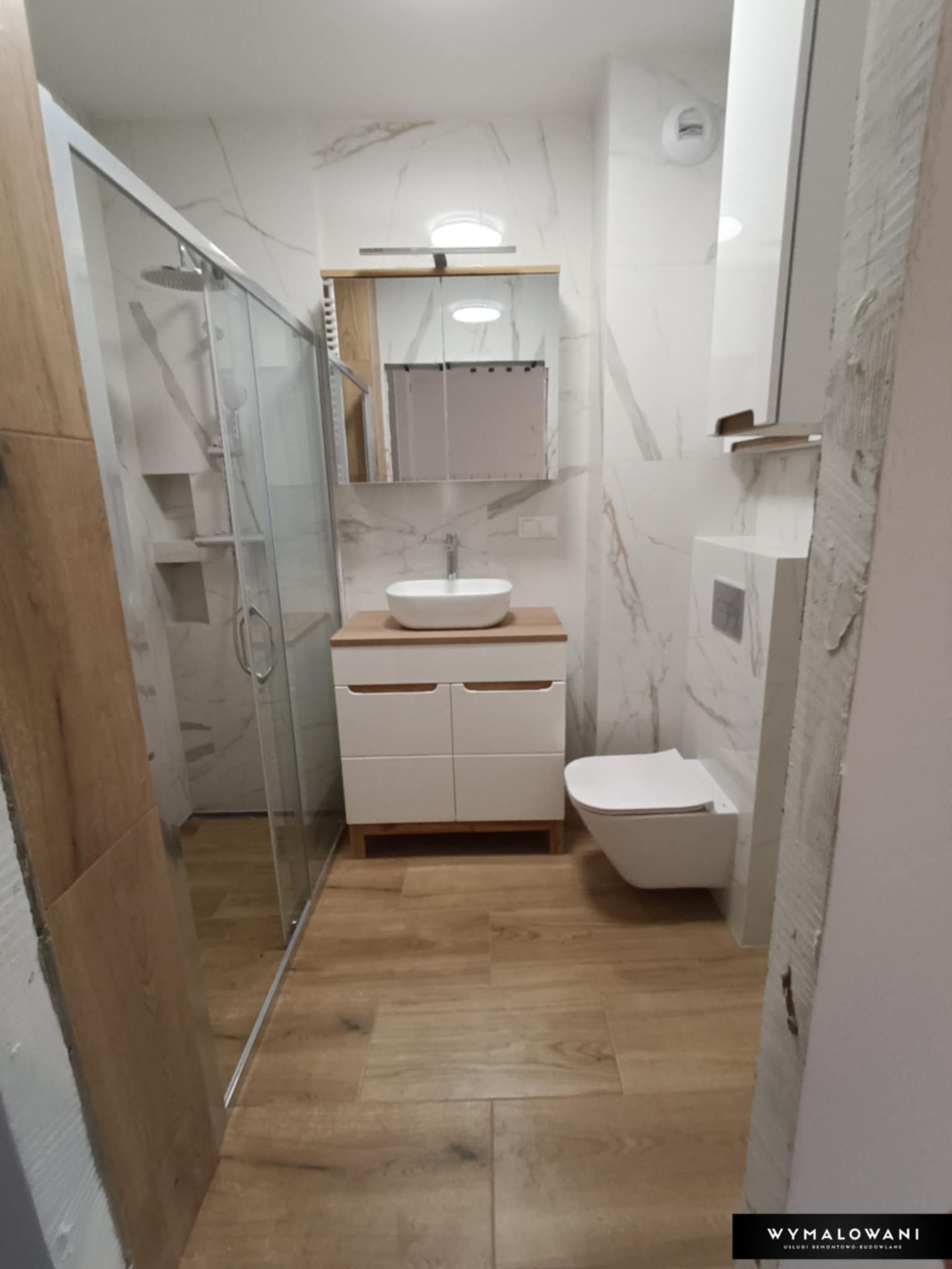 Wykończona łazienka z kabiną prysznicową, umywalką nablatową na szafce z drewnianym blatem, podwieszaną toaletą i płytkami imitującymi marmur na ścianach, oraz drewnopodobną podłogą
