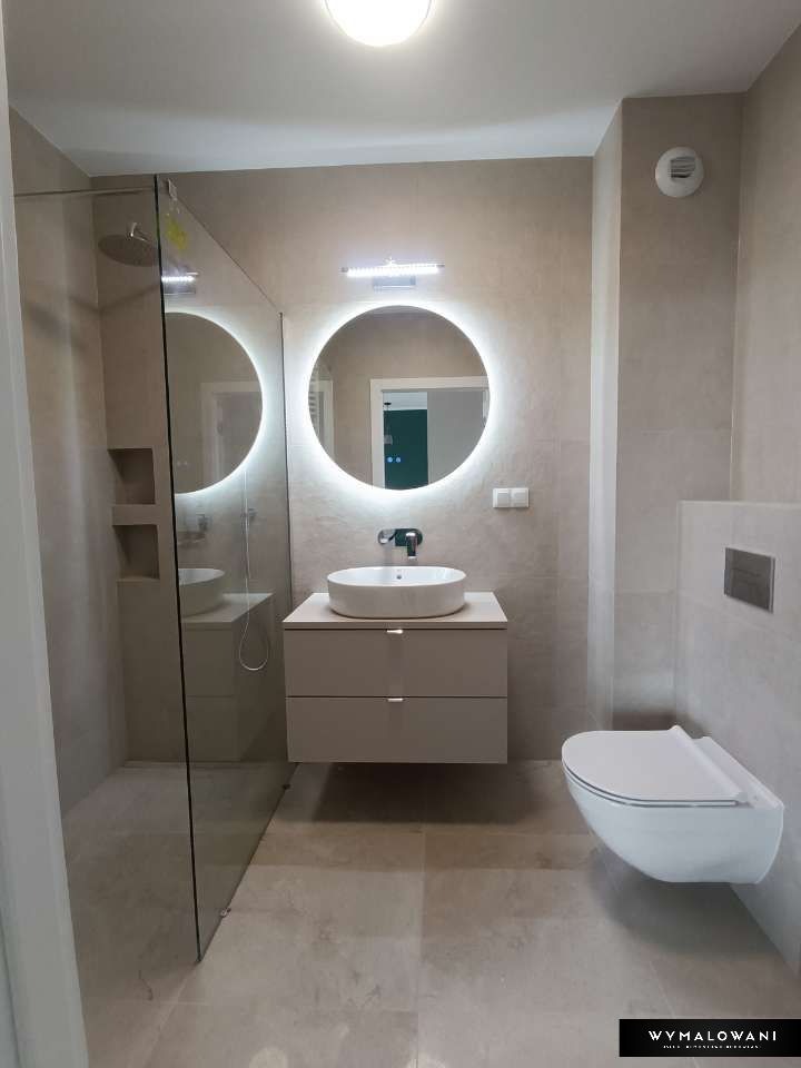 Nowoczesna łazienka w beżowych odcieniach z okrągłym lustrem podświetlanym LED, wiszącą toaletą i kabiną prysznicową typu walk-in z widoczną deszczownicą.
