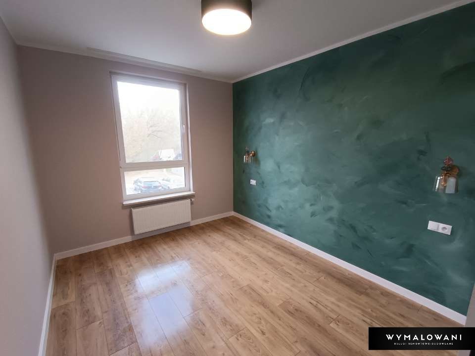 Pokój po remoncie z drewnopodobną podłogą, ścianą w odcieniach zieleni z dekoracyjnym efektem, oświetleniem ściennym i grzejnikiem pod oknem.