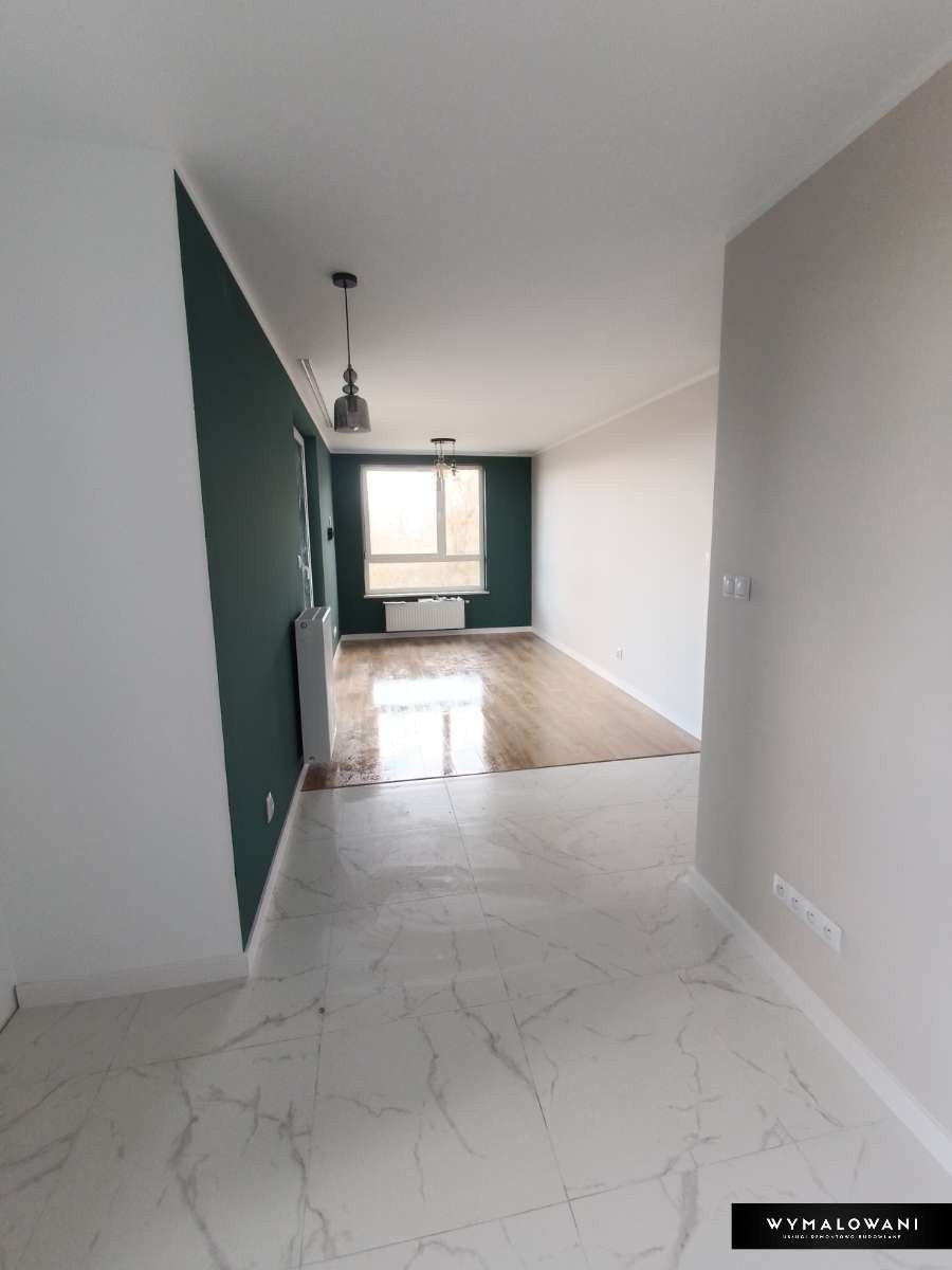 Wyremontowane wnętrze mieszkania z marmurową podłogą, ciemnozieloną ścianą i drewnianą podłogą w dalszej części pomieszczenia, widoczne wiszące lampy i biały grzejnik pod oknem.