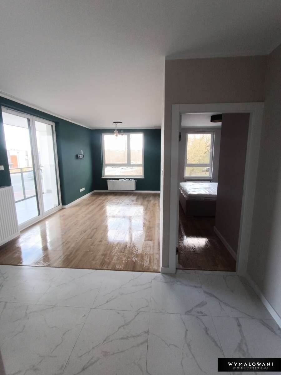 Salon po remoncie z podłogą z paneli i marmurowymi płytkami, widok na sypialnię przez otwarte drzwi, ściany w odcieniach zieleni i różu.