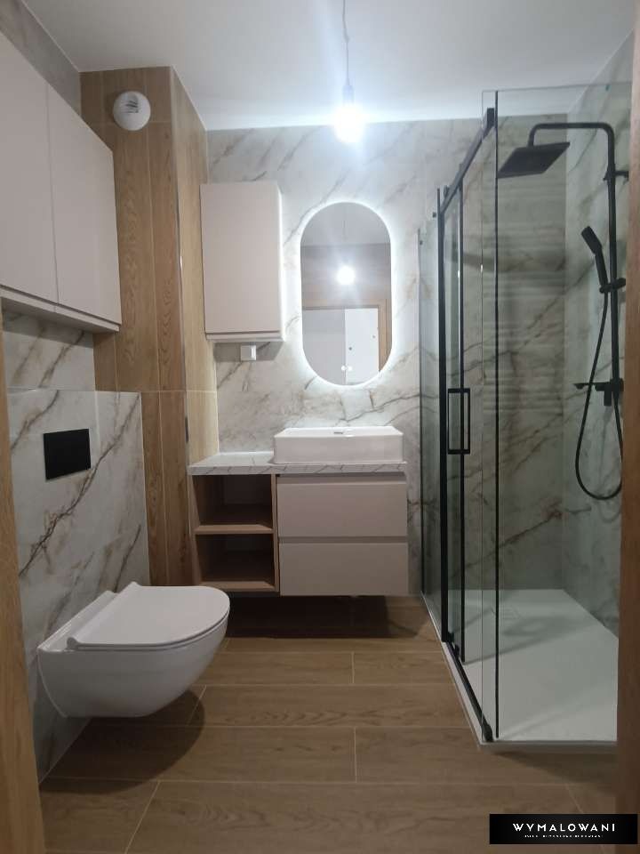 Nowoczesna łazienka z marmurowymi płytkami, podwieszaną toaletą, szafką z umywalką i podświetlanym owalnym lustrem, kabina prysznicowa z czarnymi ramami.