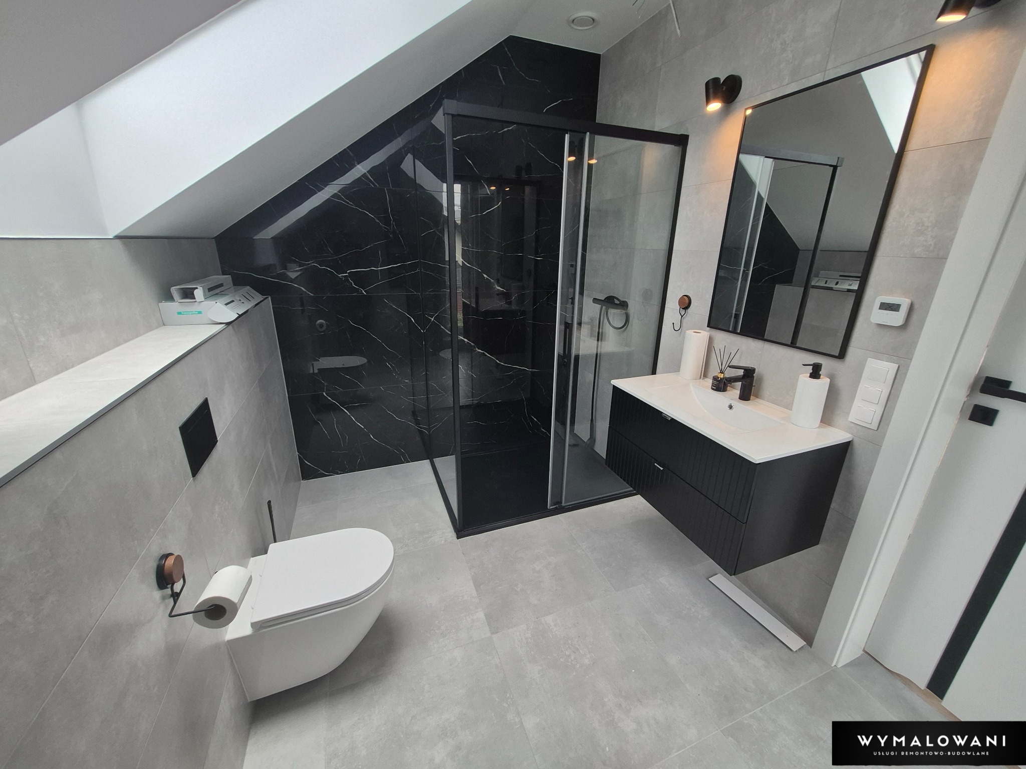 Nowoczesna łazienka na poddaszu z kabiną prysznicową wyłożoną czarnym marmurem, umywalką na szafce i wiszącą toaletą. Szare płytki na ścianach i podłodze.