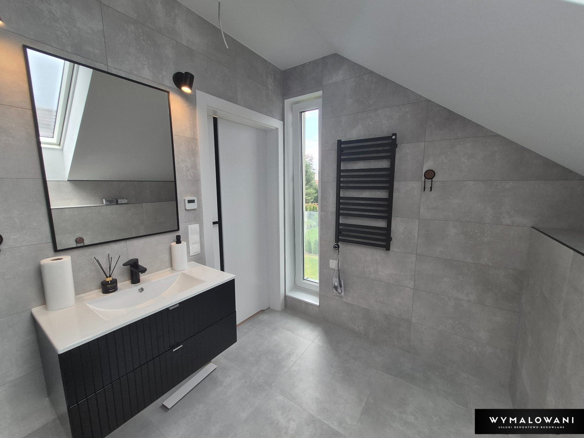 Jasnoszara łazienka na poddaszu z czarnymi akcentami: szafka pod umywalką, grzejnik i rama lustra. Okno doświetla wnętrze, widoczny skos dachu.