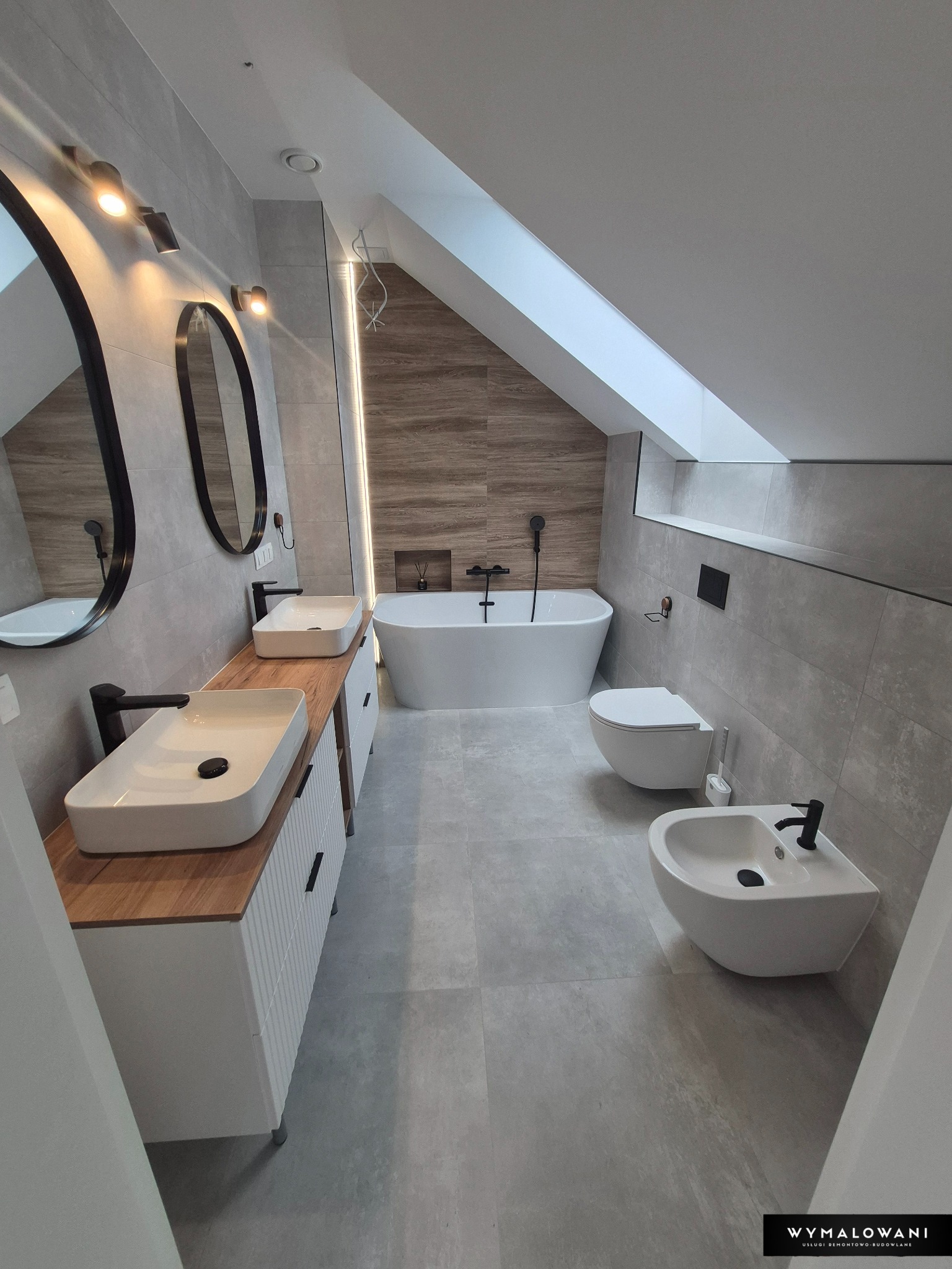 Nowoczesna łazienka na poddaszu z drewnianymi akcentami: dwie umywalki, owalne lustra, wolnostojąca wanna i bidet. Szarość płytek kontrastuje z ciepłem drewna.