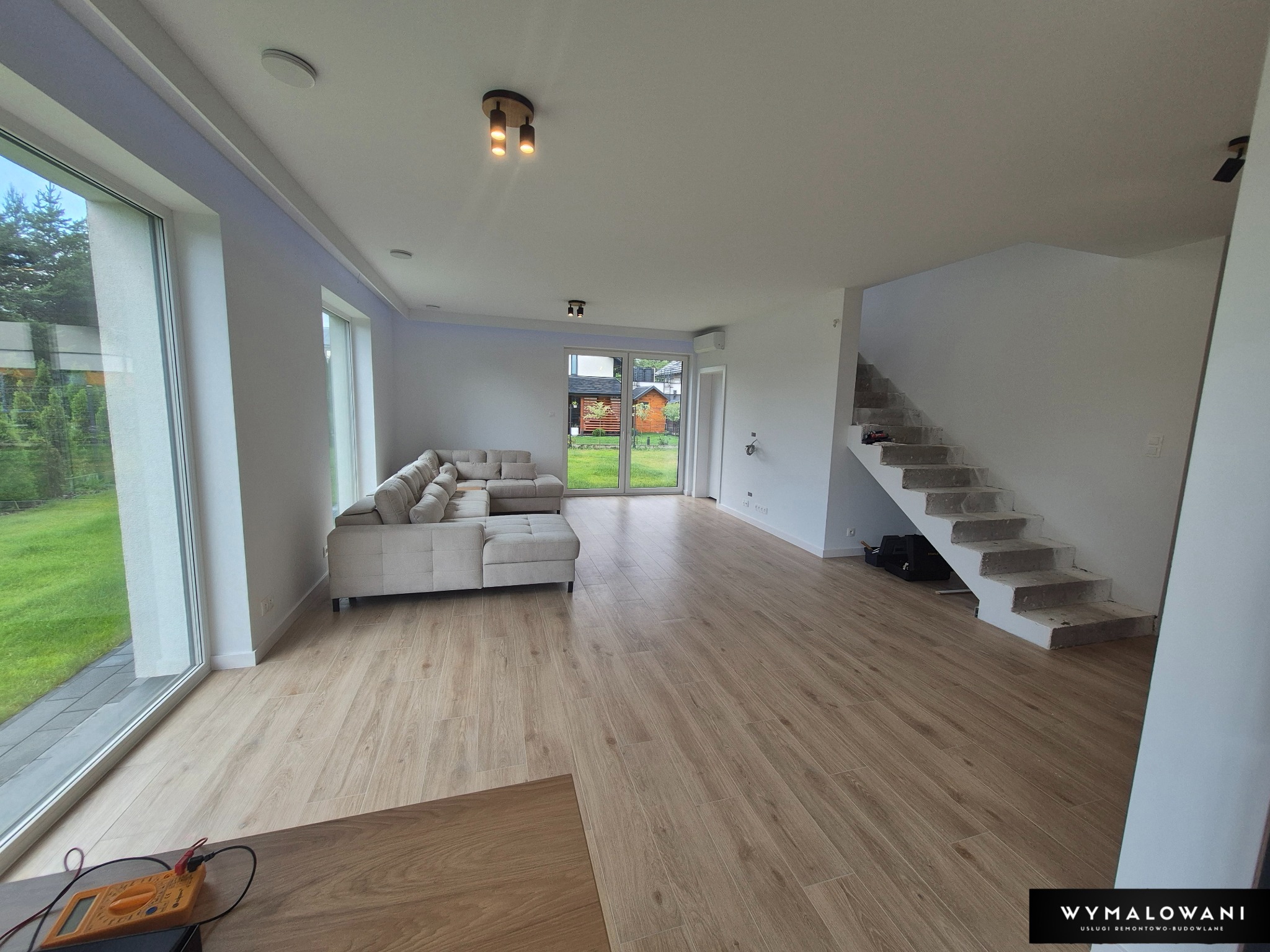 Jasne, przestronne wnętrze po remoncie: panele podłogowe, pomalowane ściany na biało, surowe schody betonowe, sofa narożna i duże okna z widokiem na zieleń.