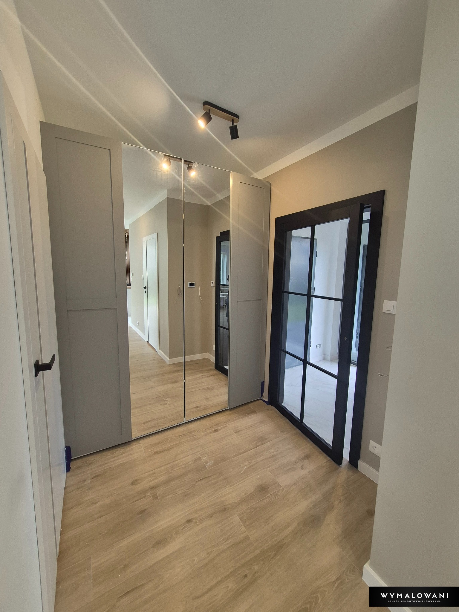Korytarz po remoncie z lustrzaną szafą wnękową i czarnymi drzwiami z przeszkleniami. Jasne panele podłogowe i stonowana kolorystyka ścian. Nowoczesne oświetlenie.