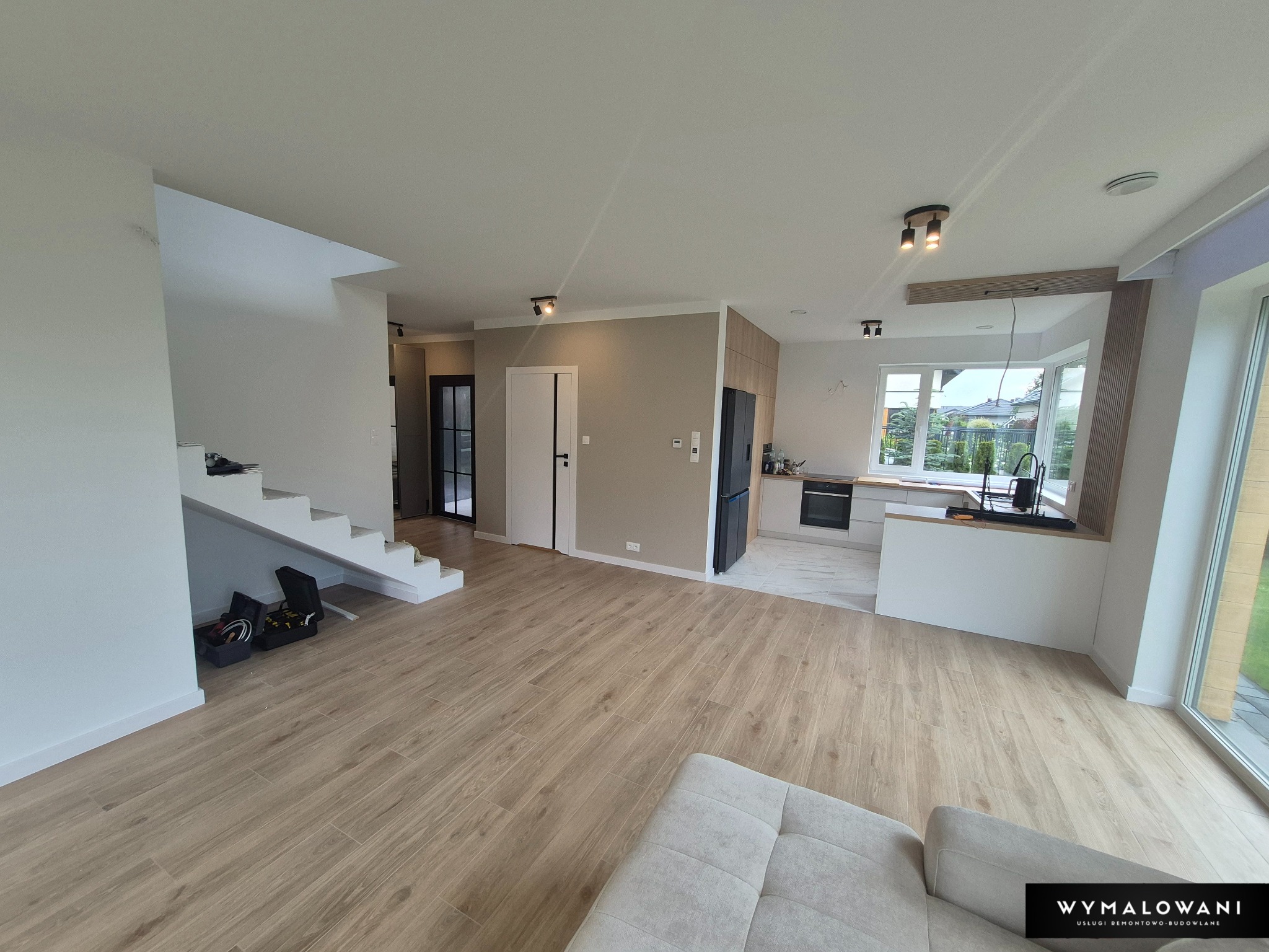 Jasne, przestronne wnętrze domu po remoncie: drewnopodobne panele, białe ściany, schody, widok na kuchnię i okno. Na podłodze leżą narzędzia.