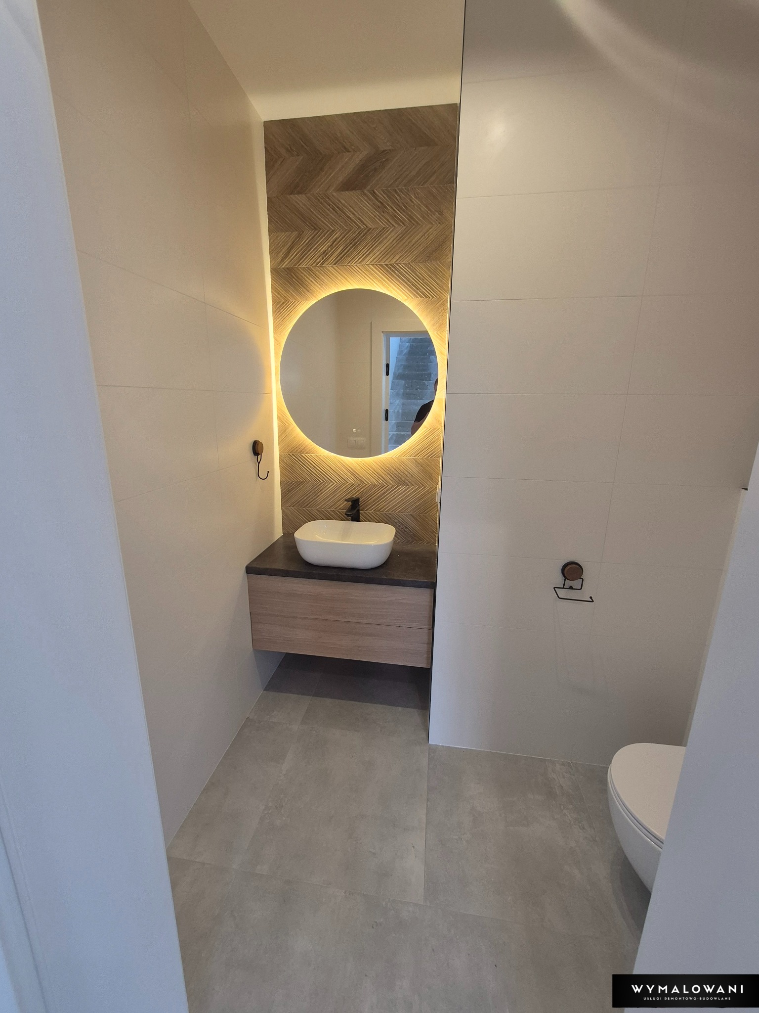 Nowoczesna łazienka z umywalką na szafce, okrągłym lustrem z podświetleniem LED i płytkami imitującymi drewno na ścianie za umywalką. Minimalistyczny design.