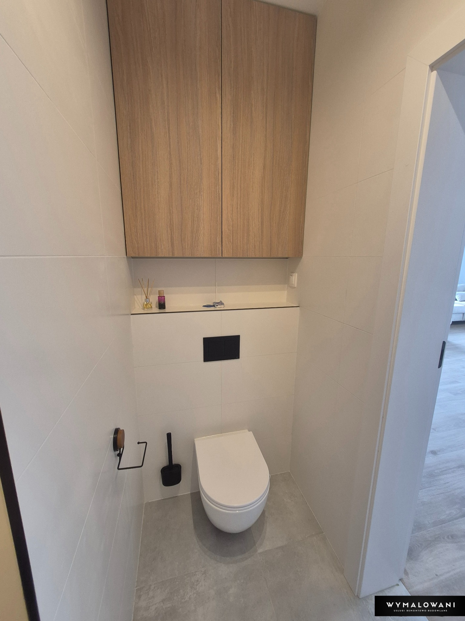 Nowoczesna toaleta po remoncie: białe płytki, drewniana szafka nad WC, szare płytki podłogowe. Minimalistyczny design, czyste linie, estetyczne wykończenie.