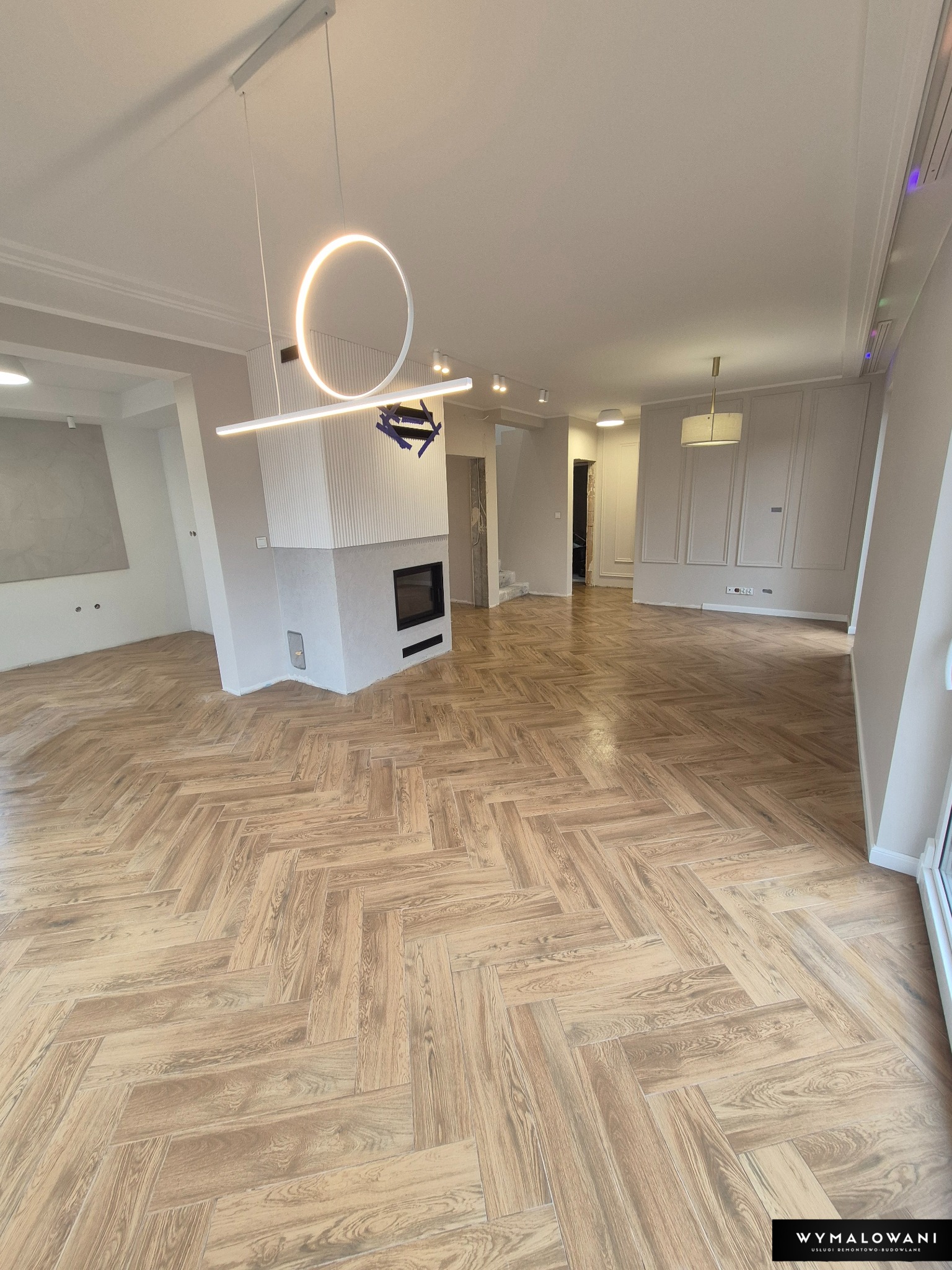 Jasne panele ułożone w jodełkę w przestronnym wnętrzu z kominkiem i nowoczesnym oświetleniem. Widoczne listwy przypodłogowe i fragmenty ścian.
