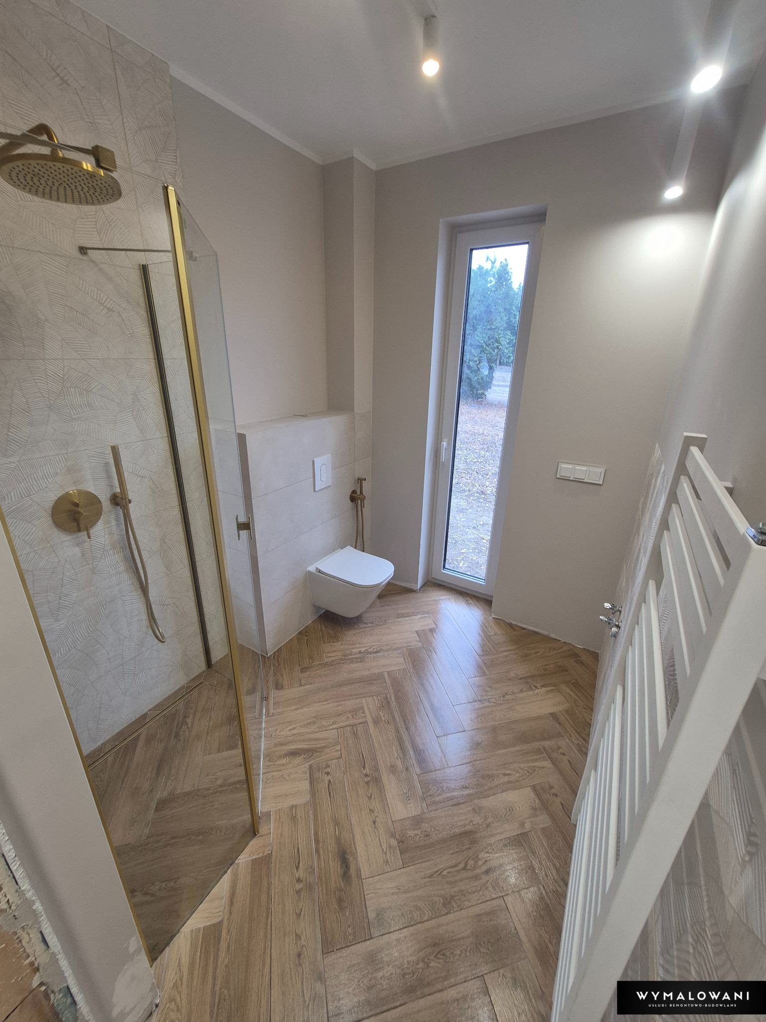 Nowoczesna łazienka po remoncie: kabina prysznicowa ze złotymi detalami, drewnopodobna podłoga ułożona w jodełkę, wisząca toaleta i grzejnik drabinkowy.