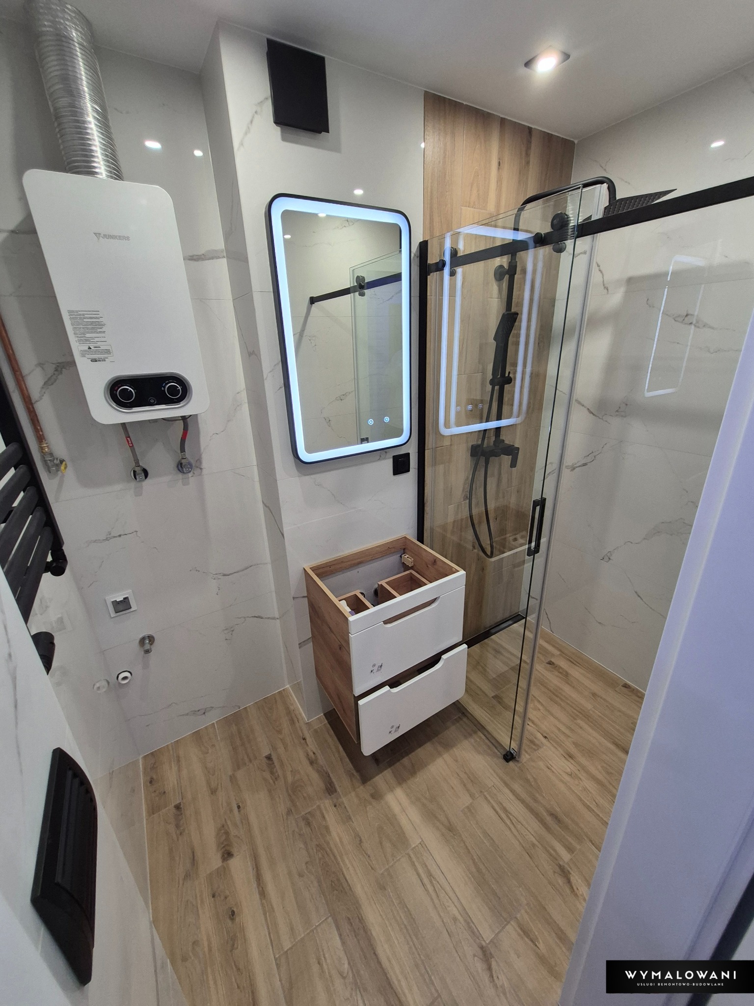 Nowoczesna łazienka po remoncie: kabina prysznicowa ze szklanymi drzwiami, podświetlane lustro, drewnopodobne płytki i biała szafka pod umywalkę z szufladami.