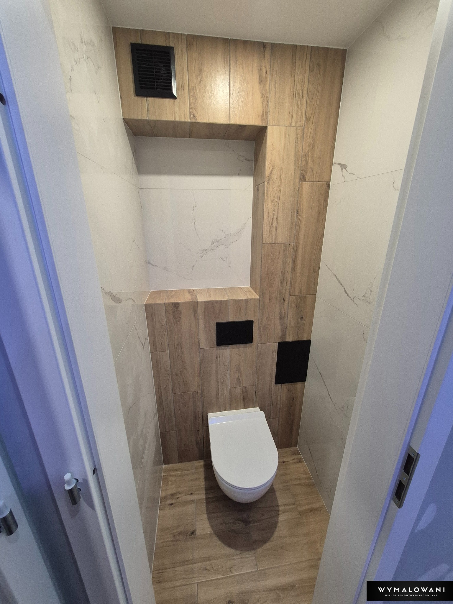 Nowoczesna toaleta z płytkami imitującymi drewno i marmur, podwieszana miska WC, czarne przyciski spłukujące, wentylator. Wykończona łazienka w neutralnych barwach.