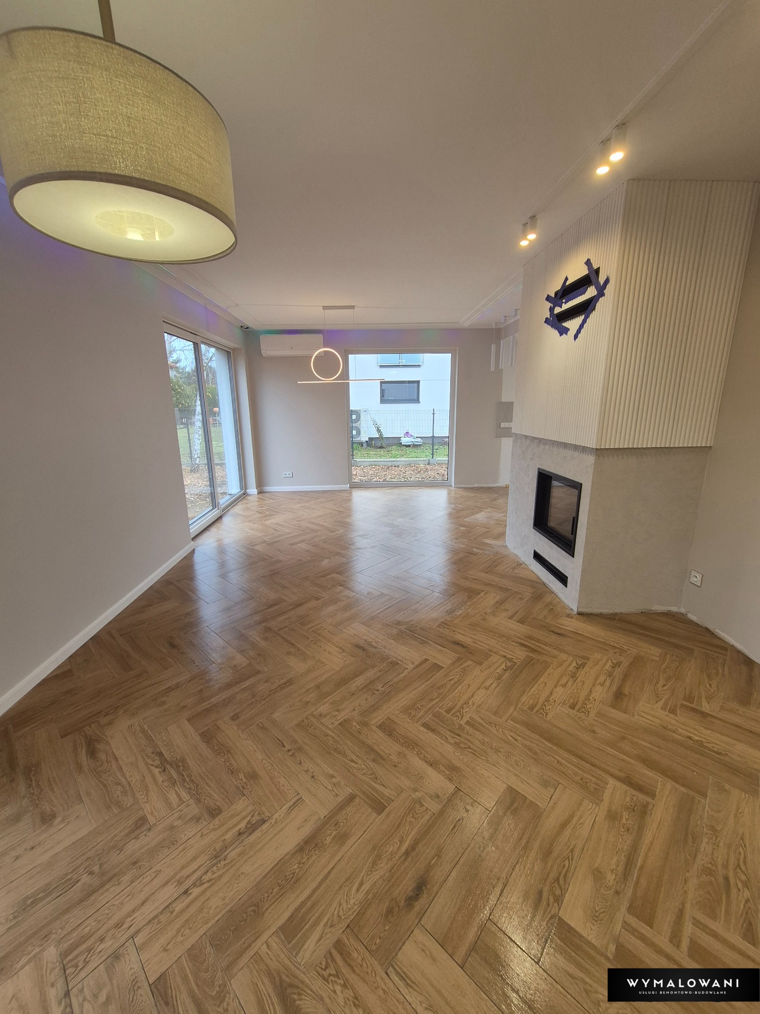 Salon po remoncie: parkiet w jodełkę, kominek z dekoracyjną ścianą, nowoczesne oświetlenie. Widok na ogród przez duże okno.