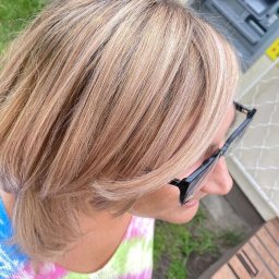 Salon Fryzjerski Paweł Zawadzki - Blond włosy z refleksami, krótko obcięte, widoczne z góry, okulary z czarnymi oprawkami na twarzy, kolorowa bluzka w tle.