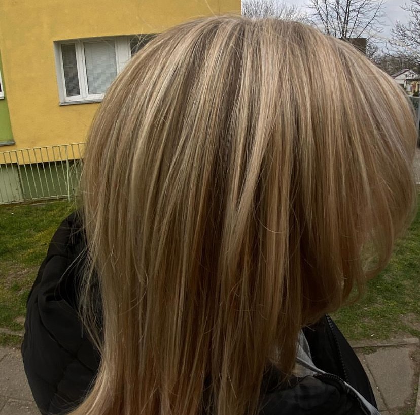 Blond włosy z refleksami w odcieniach beżu i złota, proste, sięgające ramion, na tle żółtego budynku i zieleni.