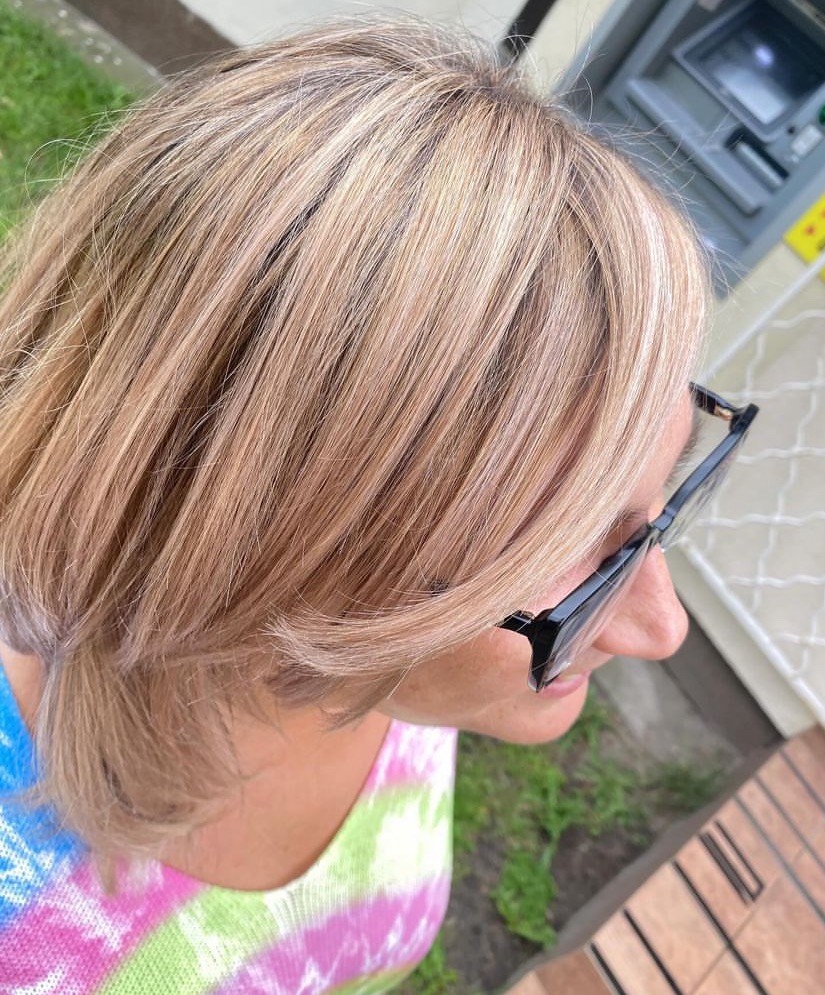 Blond włosy z refleksami, krótko obcięte, widoczne z góry, okulary z czarnymi oprawkami na twarzy, kolorowa bluzka w tle.