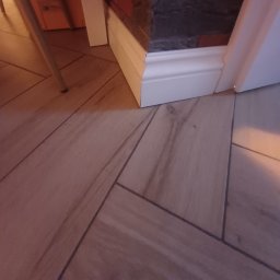 Fhu jago - Ułożona pod kątem jasna, drewnopodobna podłoga z fugami, biała listwa przypodłogowa z dekoracyjnym frezem, fragment ściany z szarej cegły i noga krzesła w rogu.