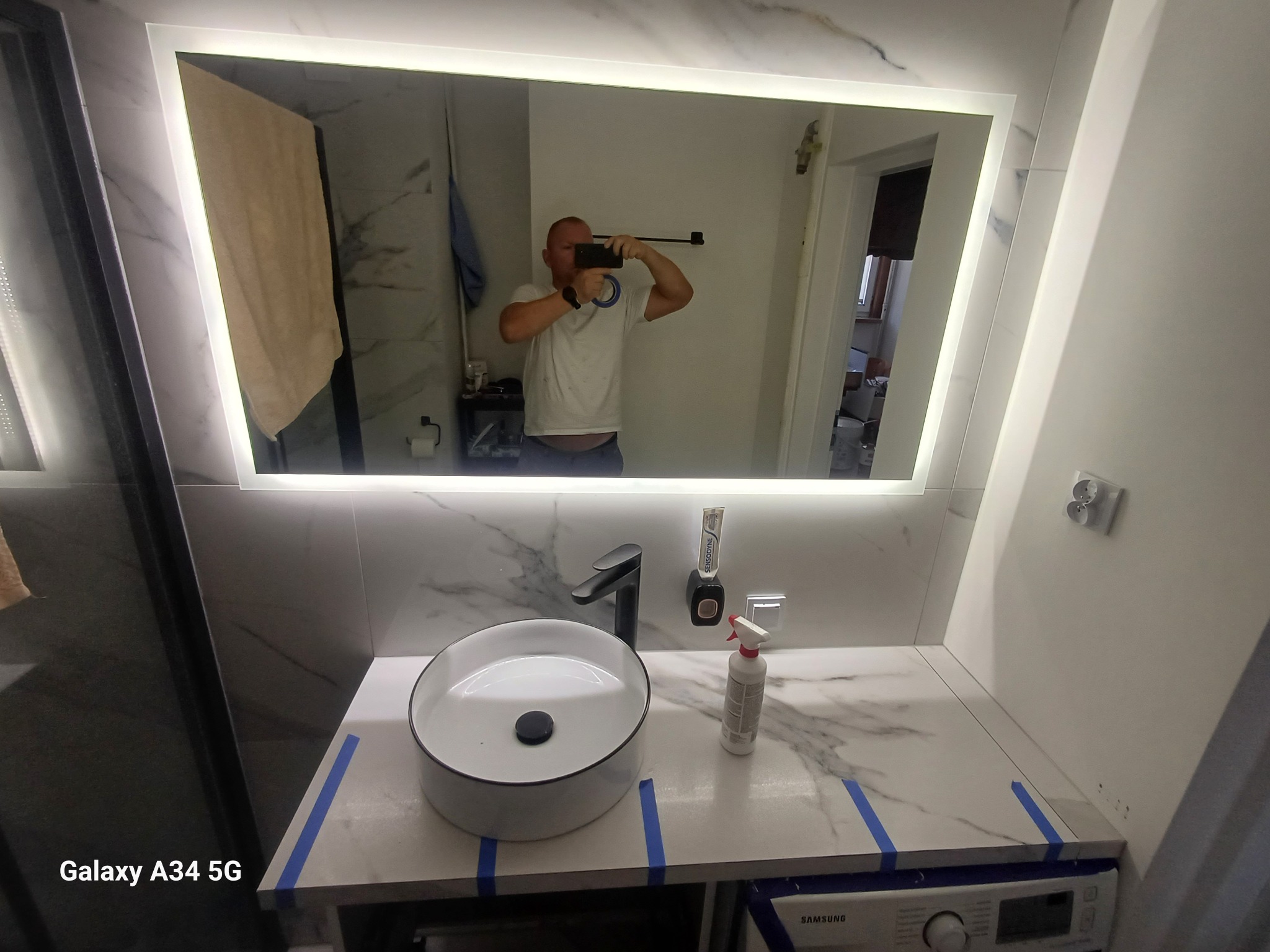 Nowoczesna łazienka z okrągłą umywalką nablatową, czarną armaturą i podświetlanym lustrem. Na blacie widoczna butelka ze spryskiwaczem i dozownik do pasty do zębów. Taśma malarska zabezpiecza blat.