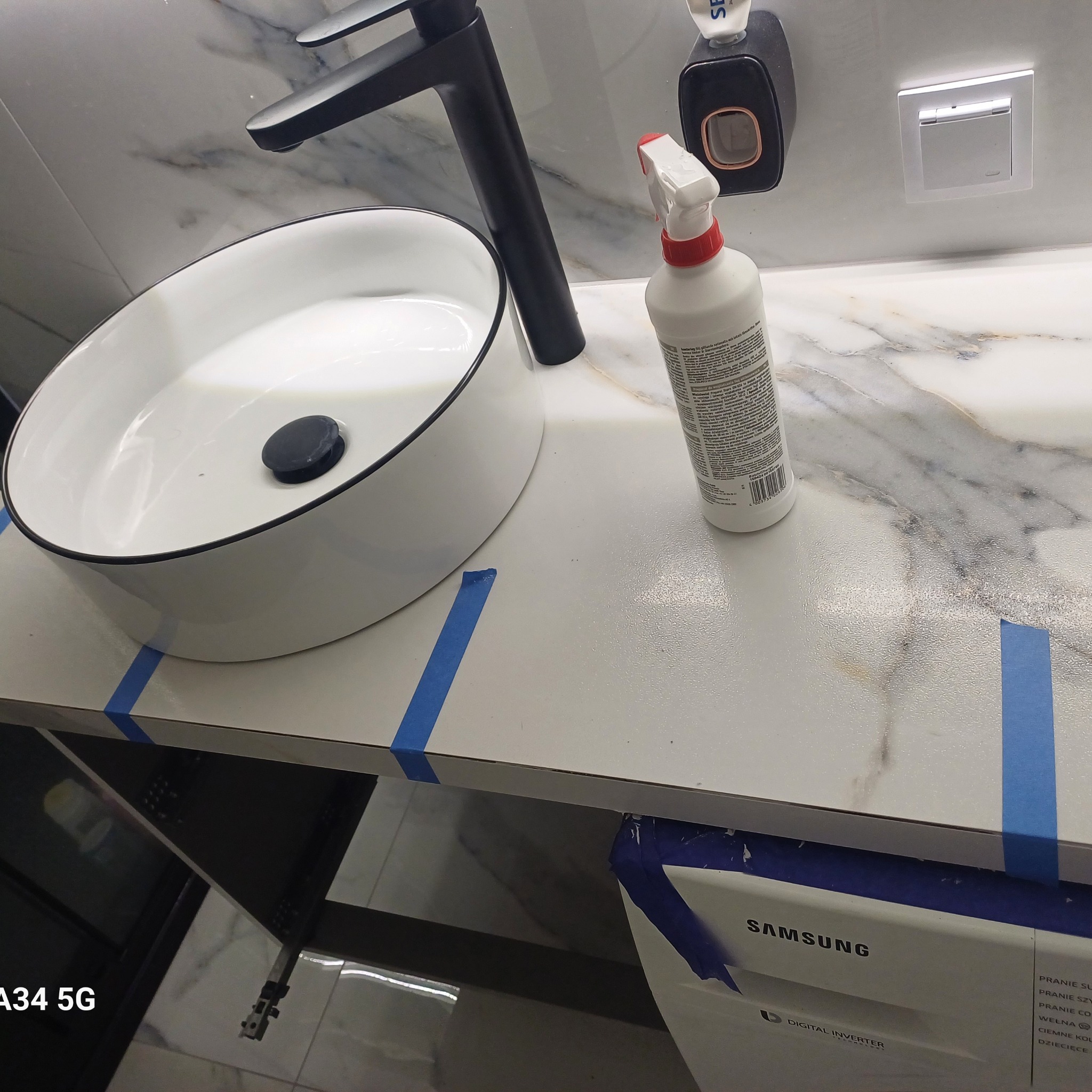 Biała umywalka nablatowa z czarną armaturą na blacie zabezpieczonym niebieską taśmą malarską, obok stoi butelka z rozpylaczem, w tle marmurowe wykończenie ściany i fragment pralki Samsung.