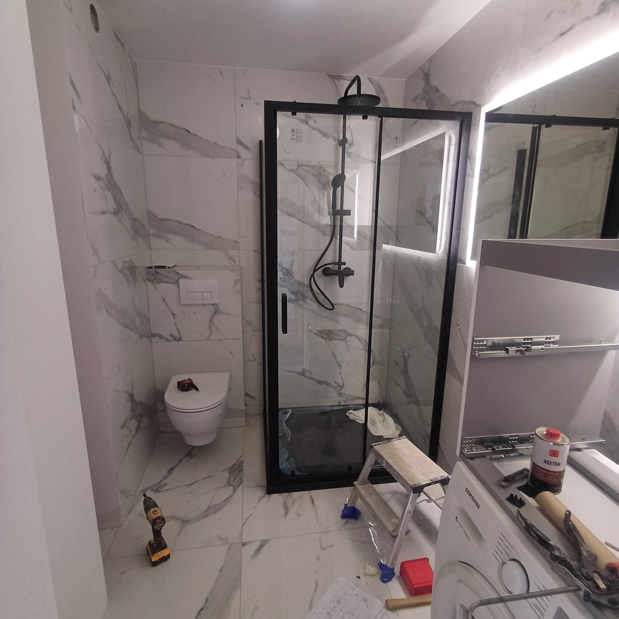 Nowoczesna łazienka w trakcie wykańczania: kabina prysznicowa z czarnymi ramami, marmurowe płytki na ścianach i podłodze, wisząca toaleta, drabina malarska, wiertarka na podłodze i pralka...