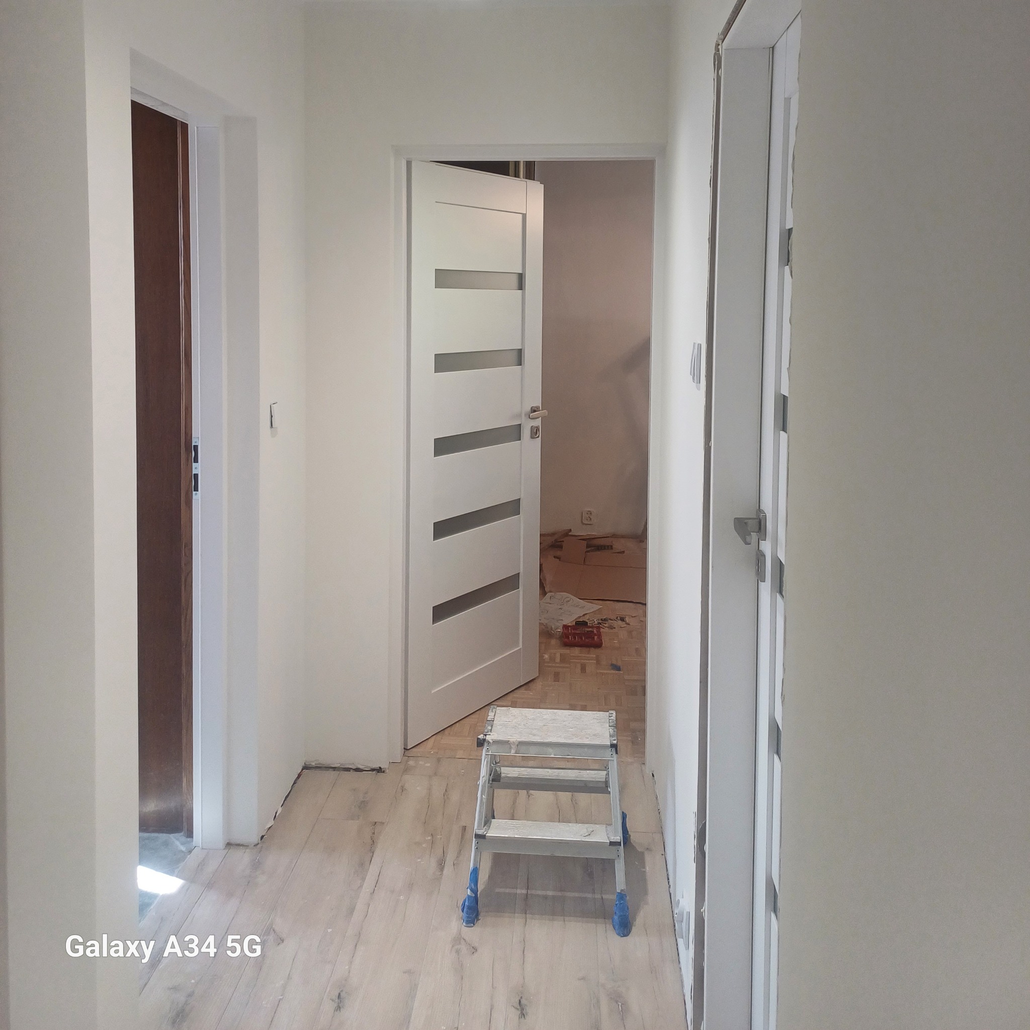 Korytarz w trakcie remontu z widocznymi drzwiami wewnętrznymi, drabinką malarską na podłodze i kartonami w tle.
