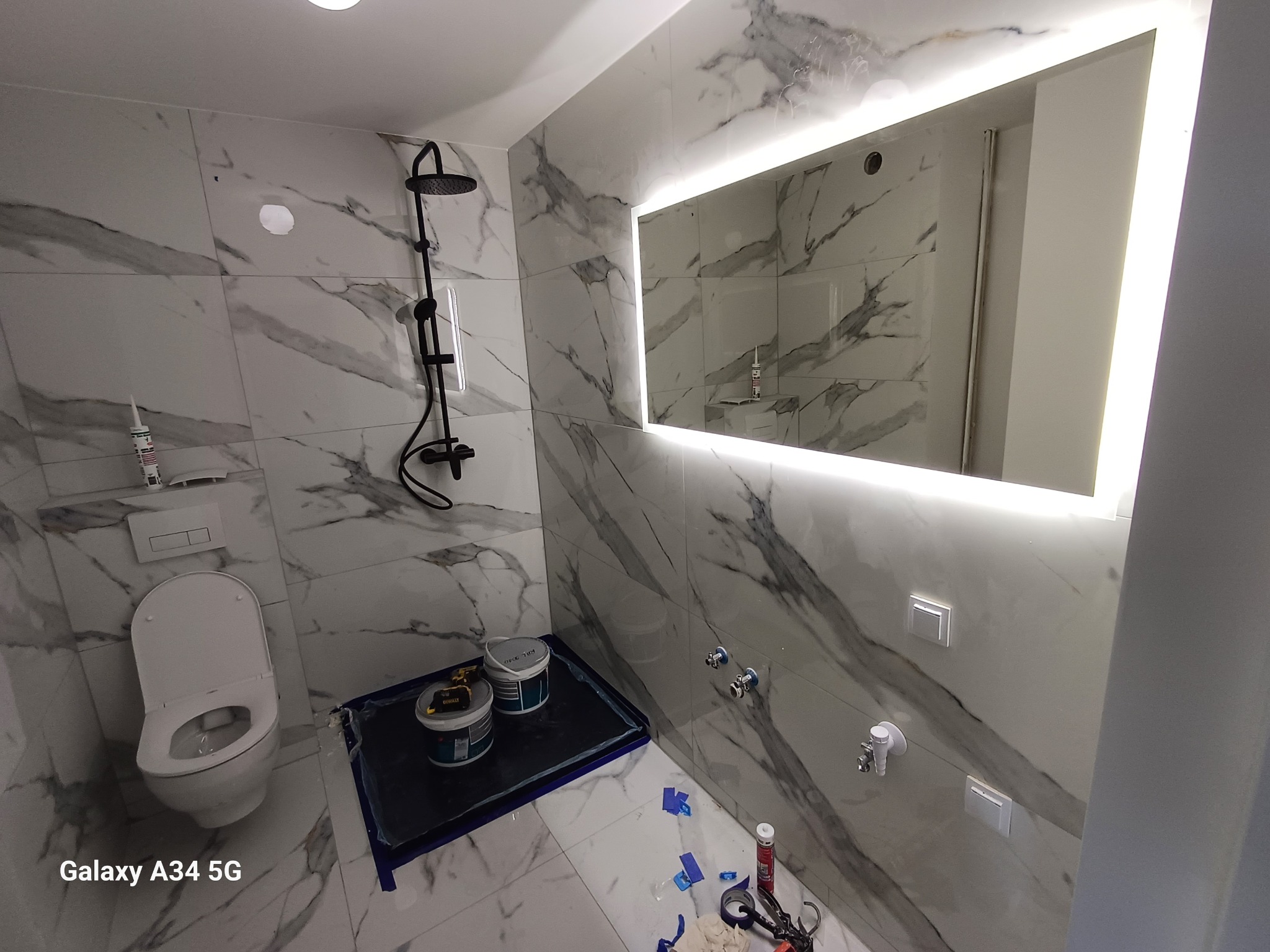 Nowoczesna łazienka w trakcie remontu: białe marmurowe płytki, czarna deszczownica, podświetlane lustro, zabezpieczona podłoga z farbami i narzędziami.