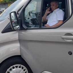 F.H.U. ROBICAR ROBERT BĄKOWSKI - Kierowca w srebrnym vanie Ford Transit, rozmawia przez telefon, otwarte drzwi przesuwne, kostka brukowa na parkingu.