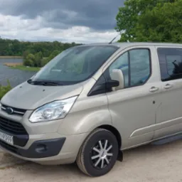 Jasny, beżowy van Ford Transit Custom z przyciemnianymi szybami, zaparkowany na szutrowej drodze z widokiem na jezioro i las w tle. Tablica rejestracyjna widoczna.