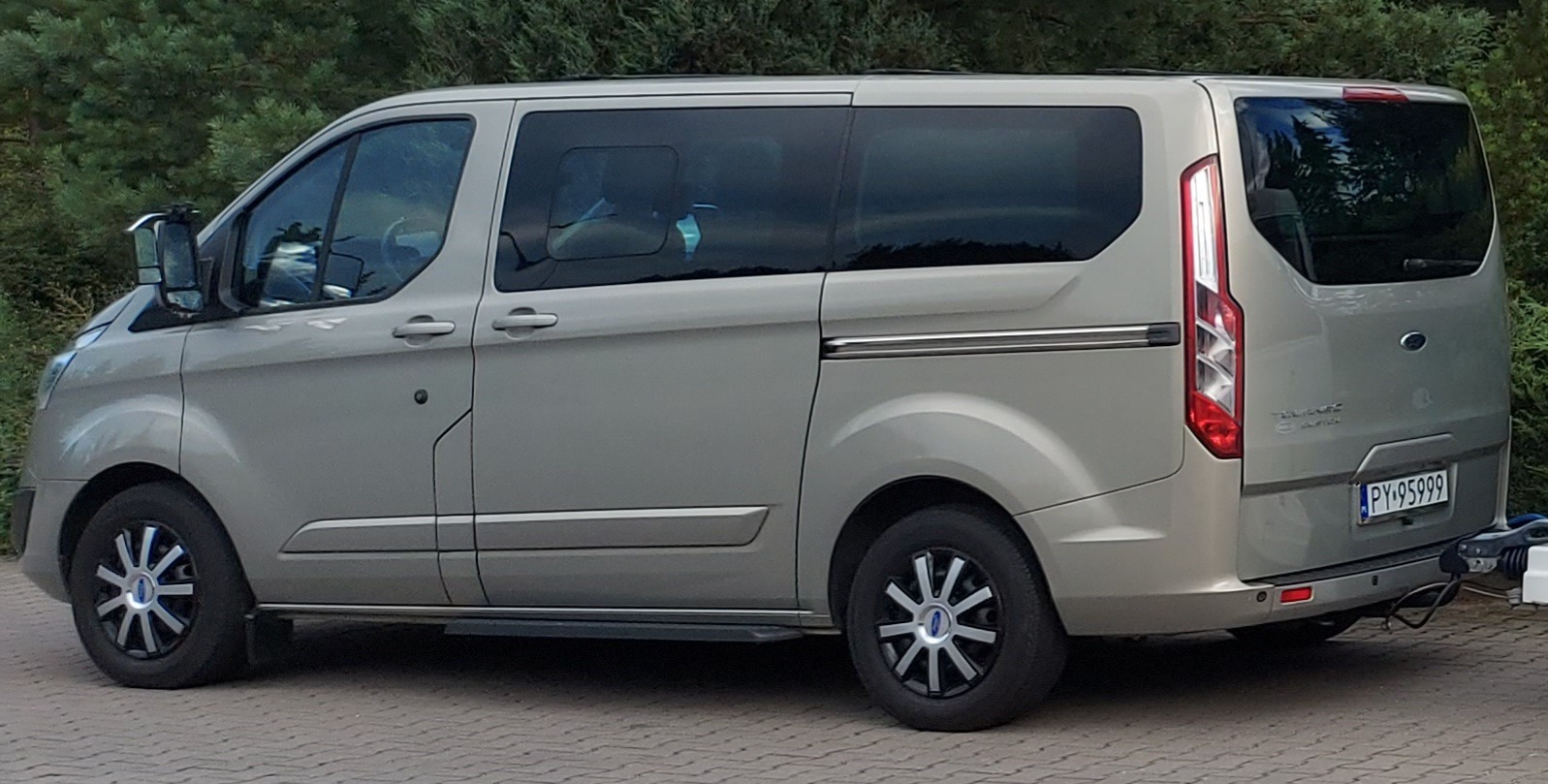 Szary Ford Transit Custom z relingami dachowymi i przyciemnianymi szybami, zaparkowany na kostce brukowej przed zielonymi drzewami.