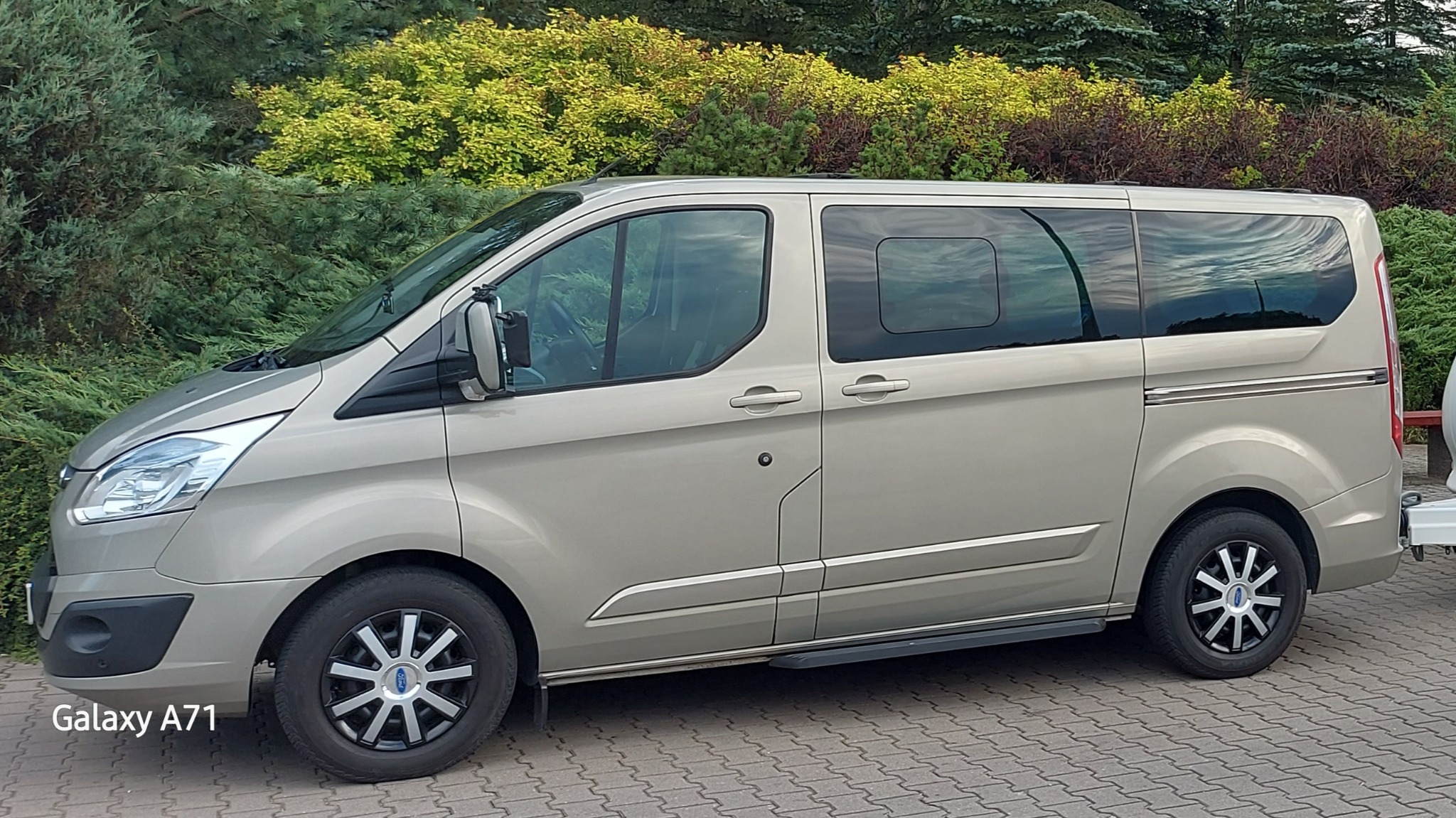 Beżowy van Ford Transit Custom z przyciemnianymi szybami i hakiem holowniczym na tle zielonych krzewów, zaparkowany na kostce brukowej.