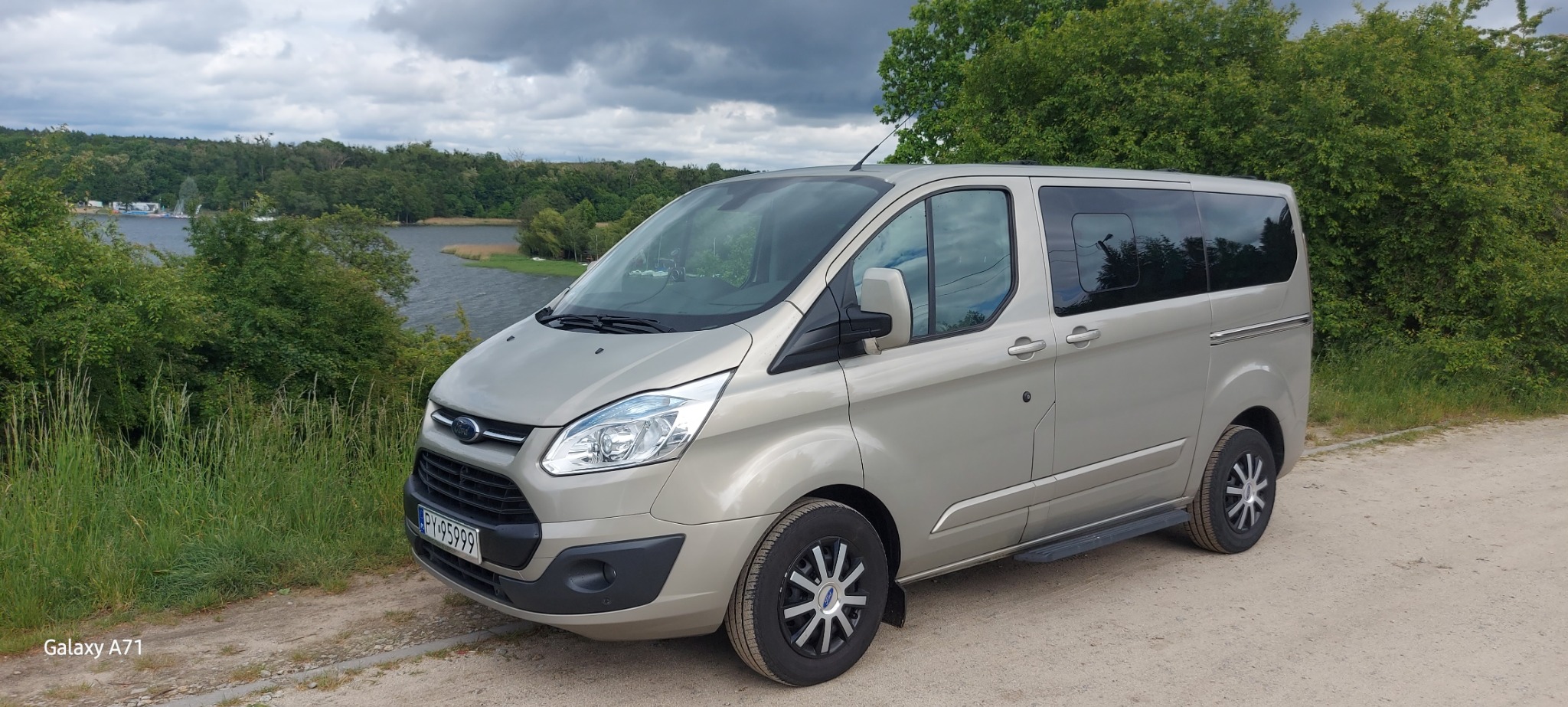 Jasny, beżowy van Ford Transit Custom z przyciemnianymi szybami, zaparkowany na szutrowej drodze z widokiem na jezioro i las w tle. Tablica rejestracyjna widoczna.