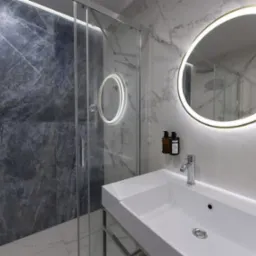 Nowoczesna łazienka z marmurowymi płytkami, okrągłym lustrem z podświetleniem LED, białą umywalką na metalowym stelażu i szklaną kabiną prysznicową.