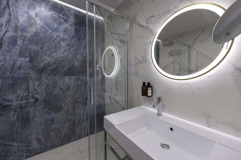 Nowoczesna łazienka z marmurowymi płytkami, okrągłym lustrem z podświetleniem LED, białą umywalką na metalowym stelażu i szklaną kabiną prysznicową.