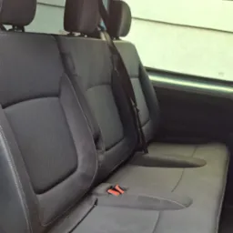 Wnętrze busa z trzema rzędami szarych foteli z zagłówkami, widok z boku, pasy bezpieczeństwa. Widoczne okno z jasnym światłem dziennym.