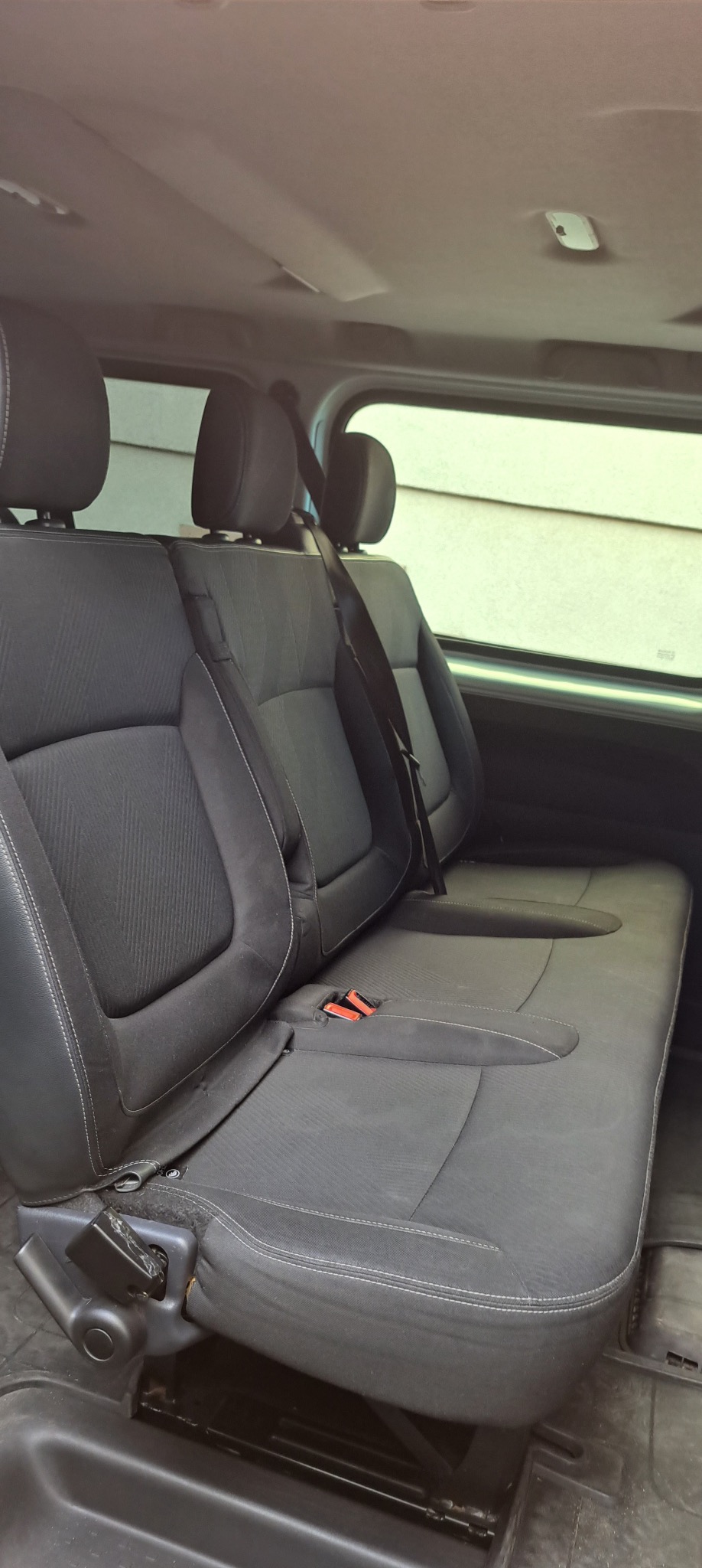 Wnętrze busa z trzema rzędami szarych foteli z zagłówkami, widok z boku, pasy bezpieczeństwa. Widoczne okno z jasnym światłem dziennym.