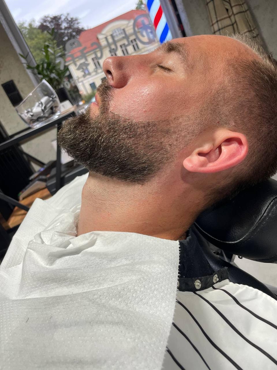 Mężczyzna z zamkniętymi oczami leży w fotelu fryzjerskim podczas trymowania brody, widoczny fragment peleryny fryzjerskiej i kubek z obciętymi włosami w tle.