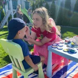 Animator w stroju wróżki maluje twarz chłopcu na tle ogrodu z domkiem zabaw.