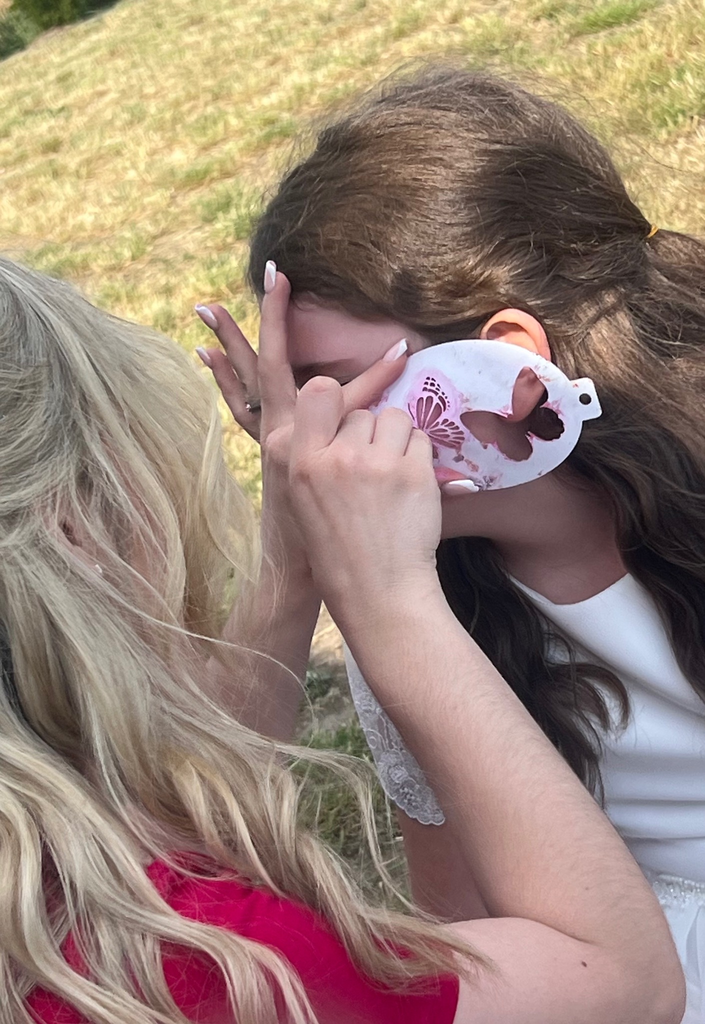 Dziewczyna z blond włosami obserwuje, jak na twarzy innej dziewczyny malowany jest wzór motyla za pomocą szablonu.