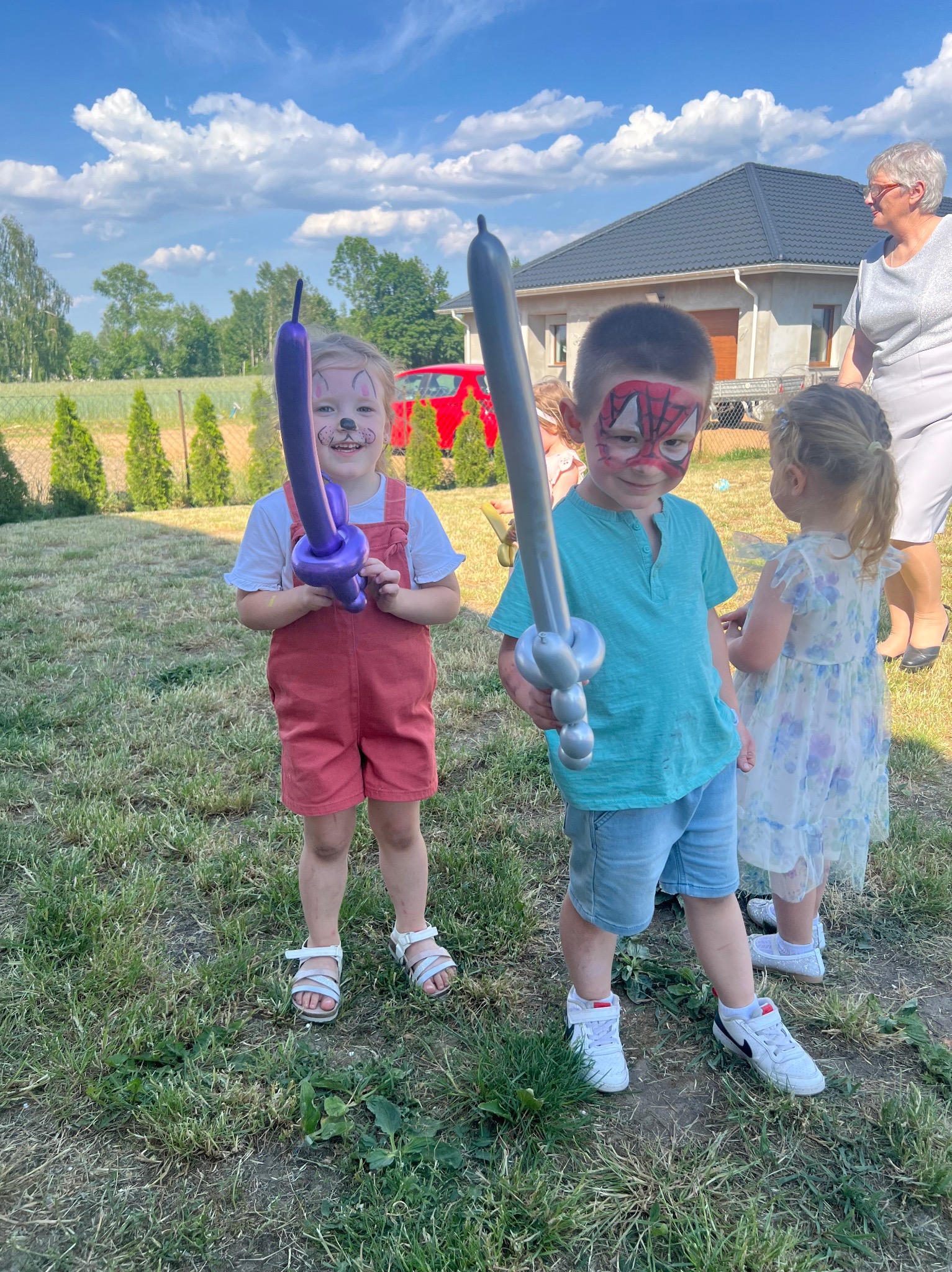 Dzieci z pomalowanymi twarzami i balonami w kształcie zwierząt i miecza, stojące na trawie przed domem podczas imprezy plenerowej w słoneczny dzień.