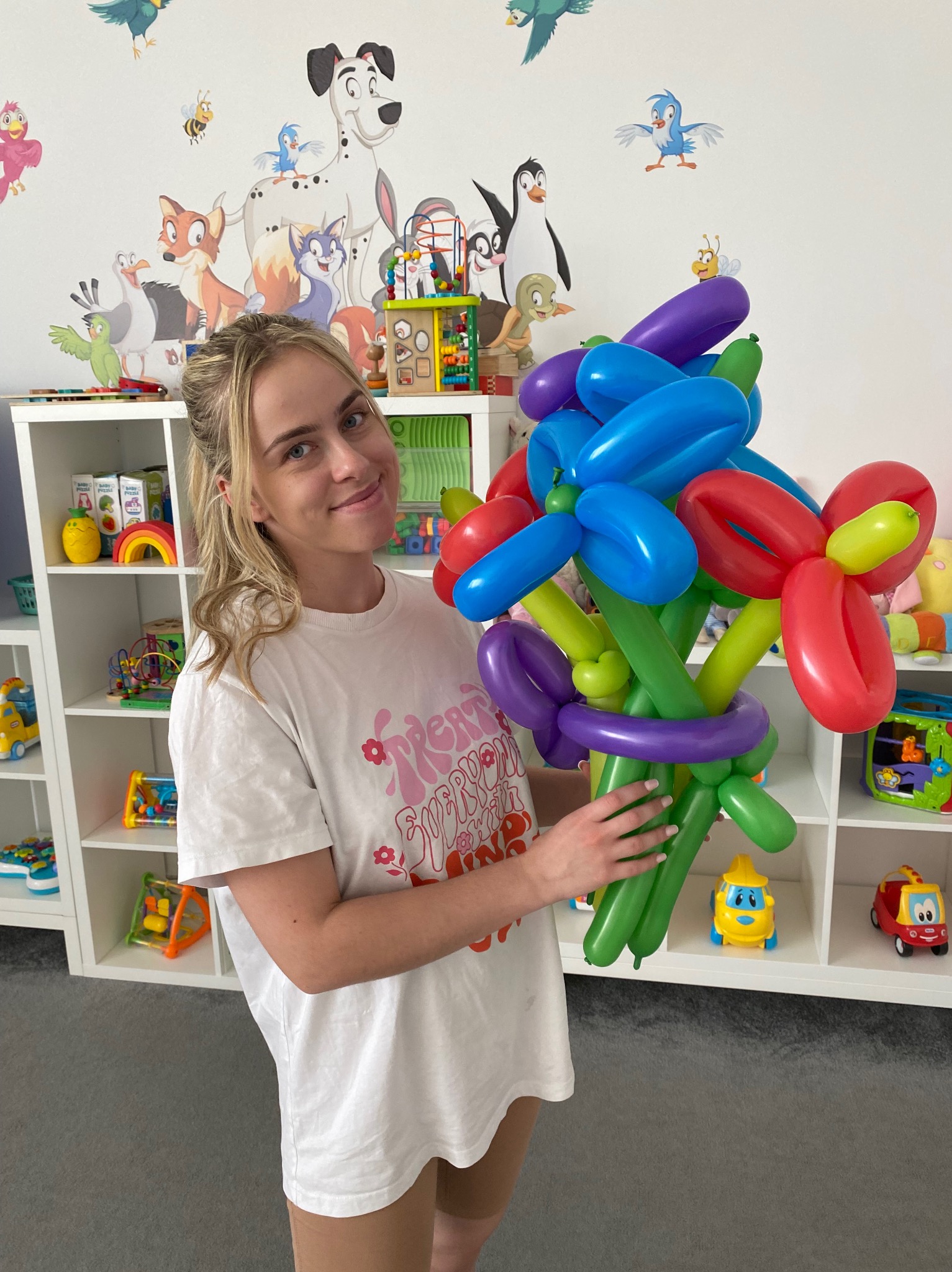 Uśmiechnięta kobieta trzyma bukiet kwiatów wykonanych z kolorowych balonów na tle pokoju dziecięcego z półkami pełnymi zabawek i ścianą ozdobioną postaciami z bajek.