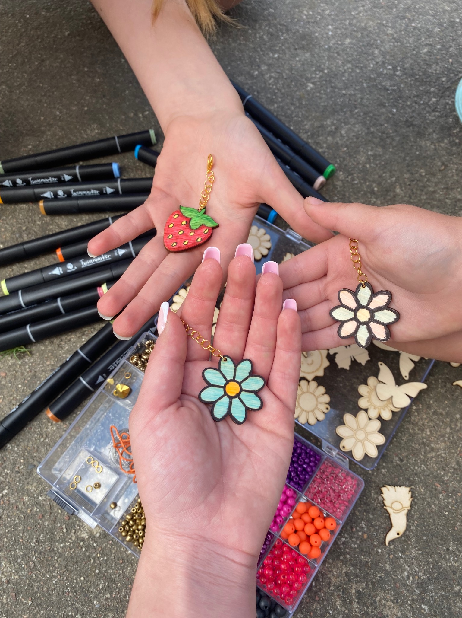 Trzy dłonie prezentują ręcznie robione, drewniane zawieszki: truskawkę i dwa kwiatki, obok markery, pudełko z koralikami i inne elementy do tworzenia biżuterii.