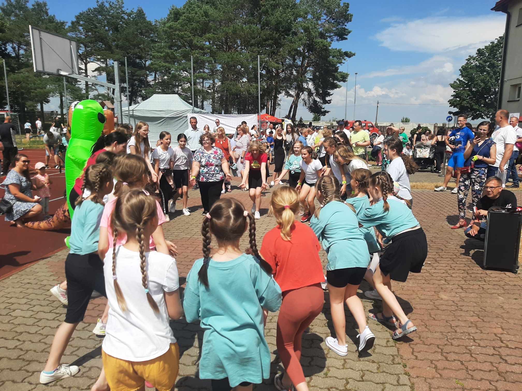 Grupa dzieci w Łomży bawi się w kółku podczas imprezy plenerowej, w tle widoczny dmuchany kostium kosmity i publiczność.