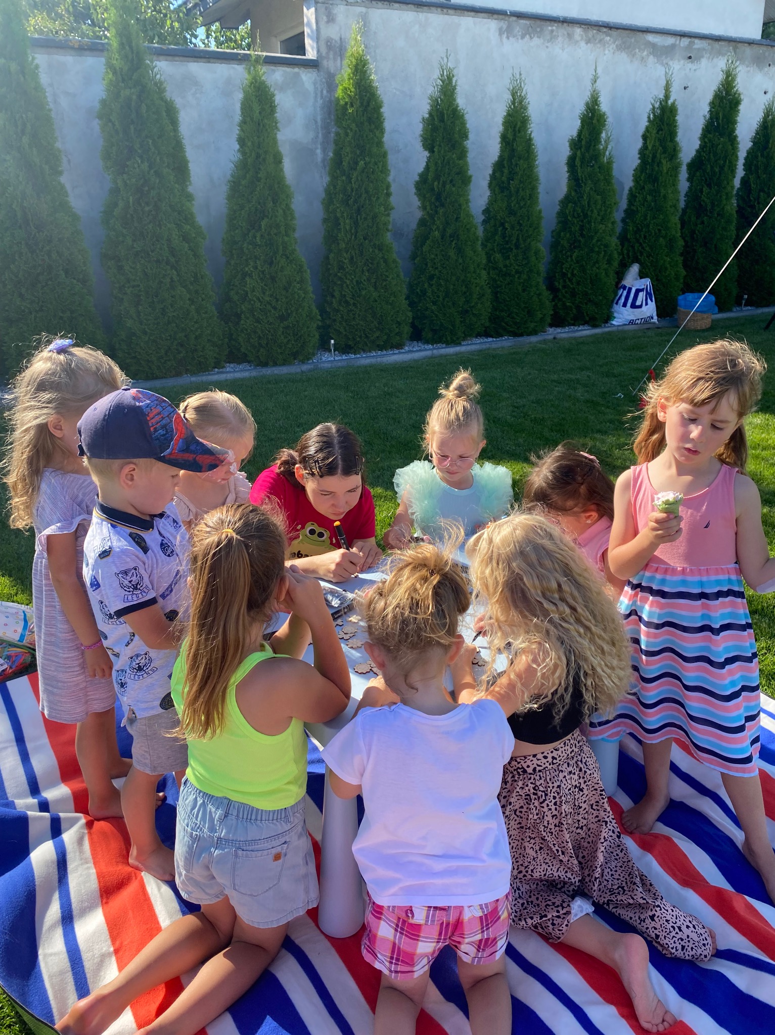 Grupa dzieci siedzi na kocu w ogrodzie, otaczając niski stolik i wykonując prace plastyczne, jedna dziewczynka trzyma loda.