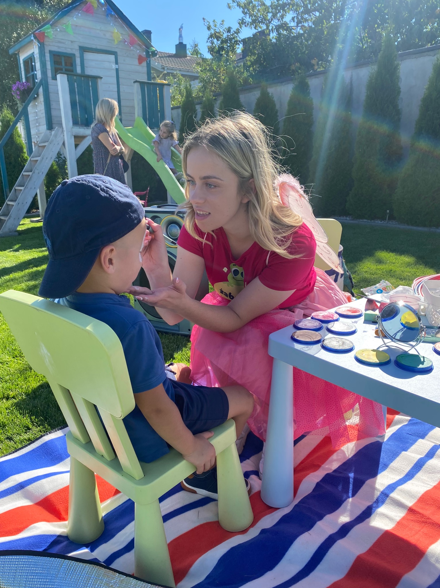 Animator w stroju wróżki maluje twarz chłopcu na tle ogrodu z domkiem zabaw.